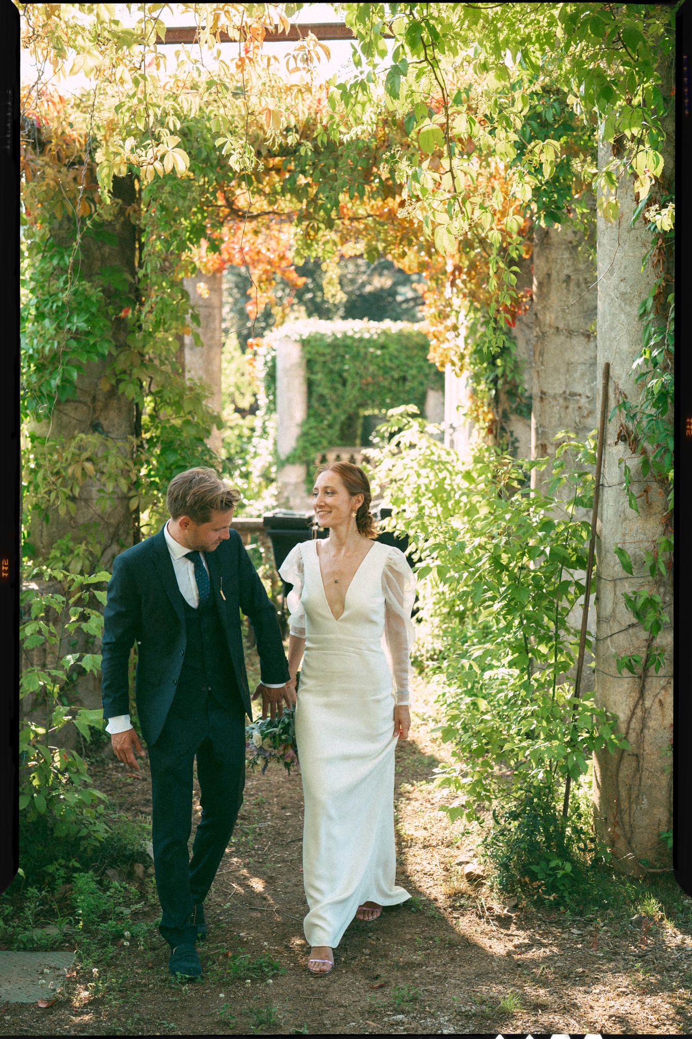 Couple walking through a sunlit garden path with trees creating a natural archway.