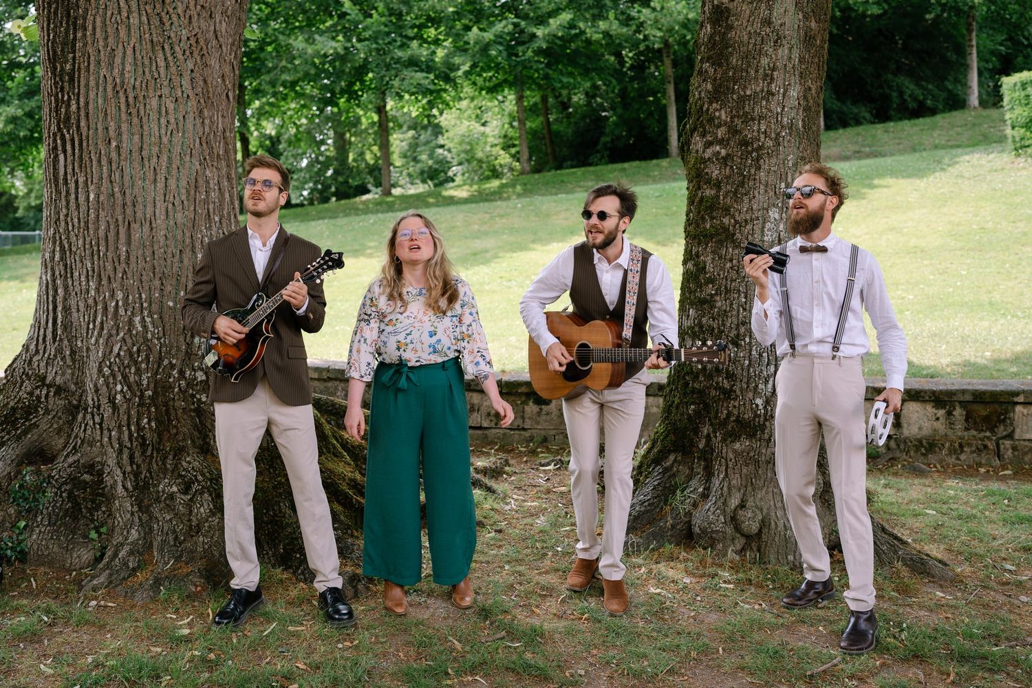 Wedding party members posing casually under large trees in a garden setting.