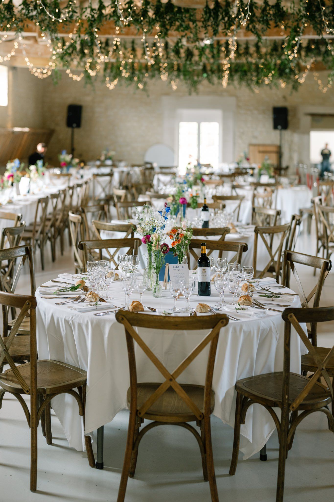 Wedding reception venue with white tablecloths, wooden chairs, and hanging greenery decorations.
