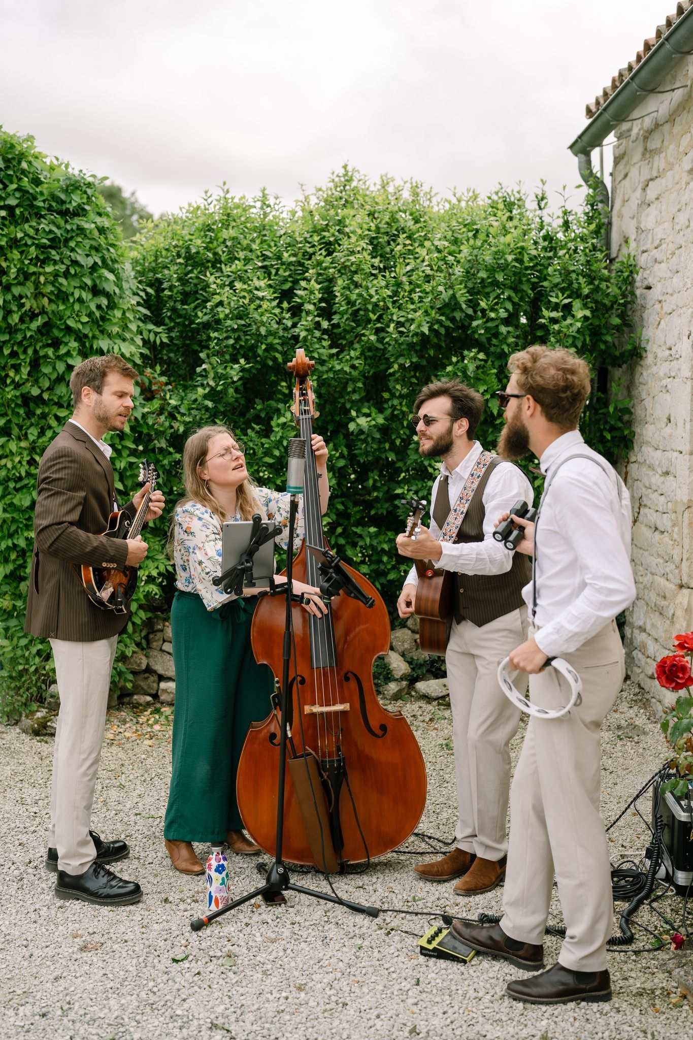 Musicians with string instruments perform outdoors against a hedge backdrop at an event.