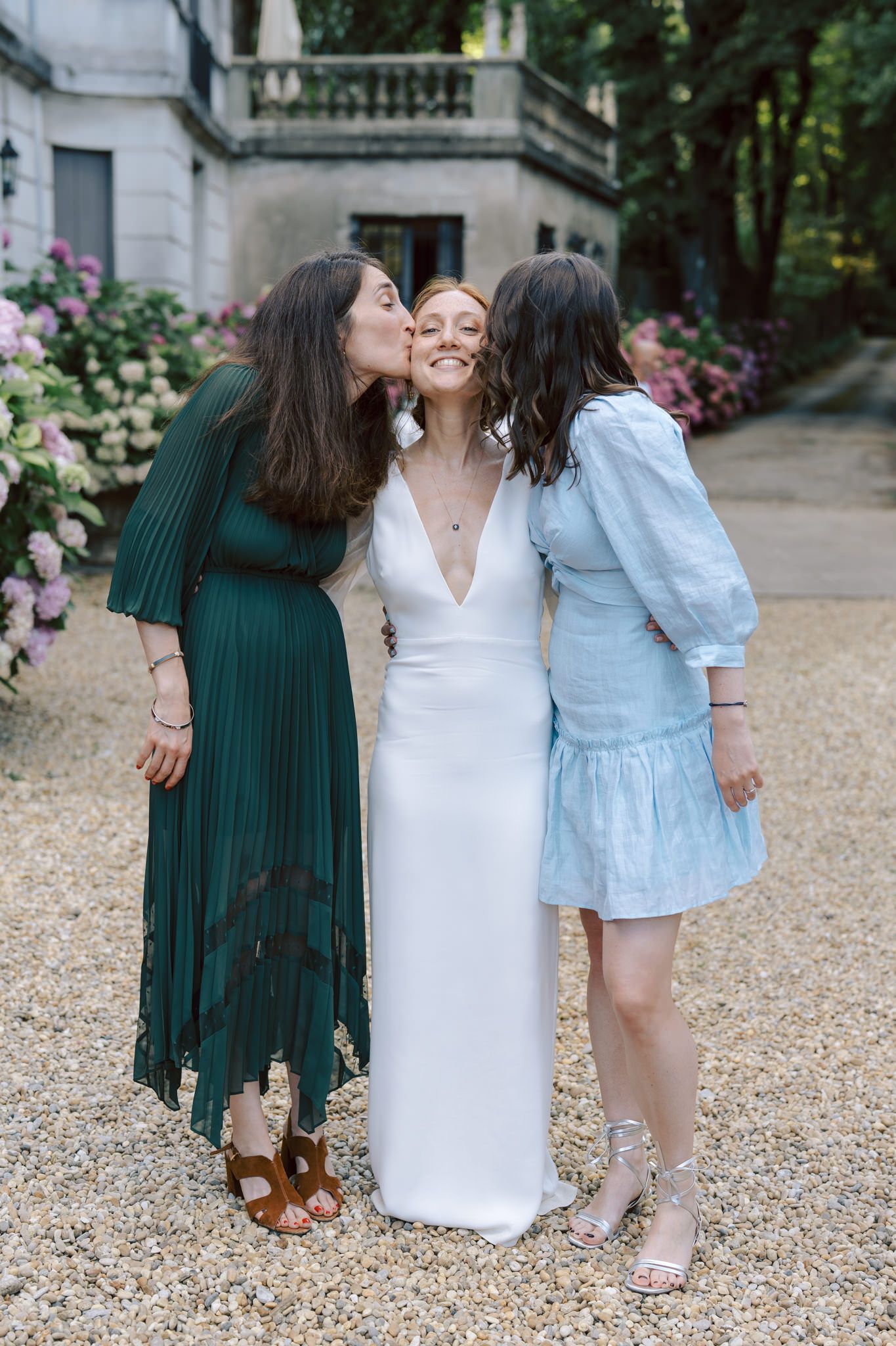 Three friends share a candid moment on a gravel path near a grand building.