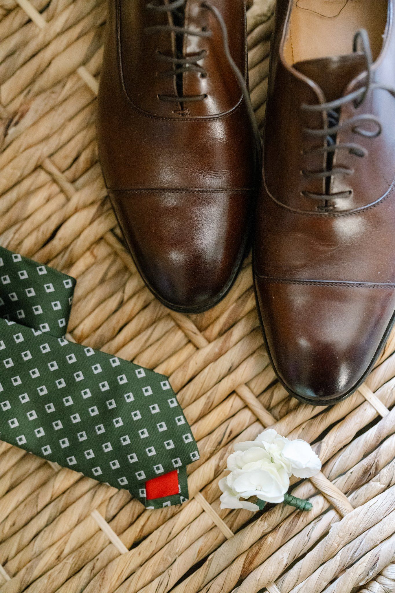 Brown leather dress shoes and green patterned tie arranged on woven surface.