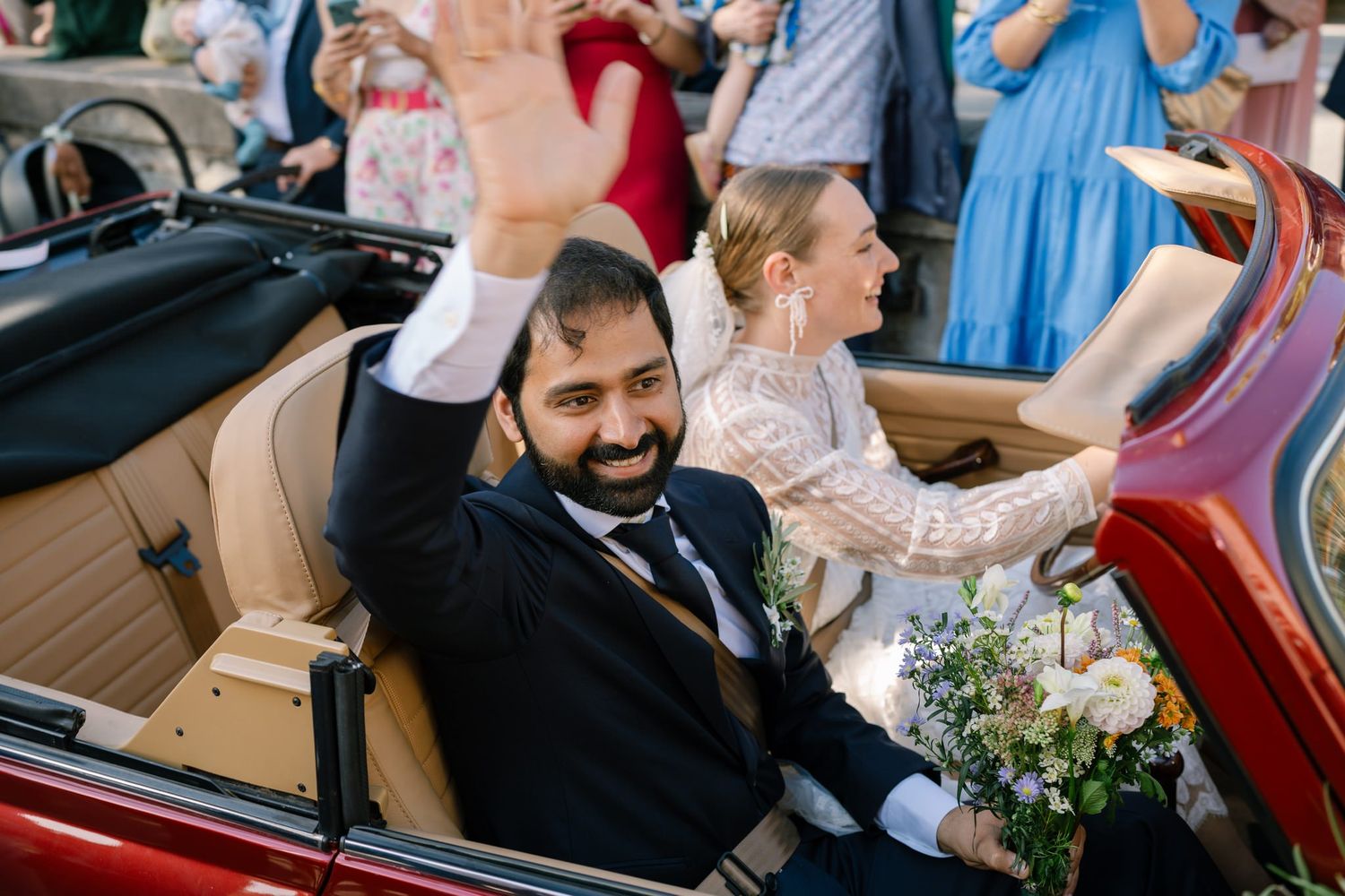 Wedding party celebrates in vintage car with floral bouquet and festive atmosphere.