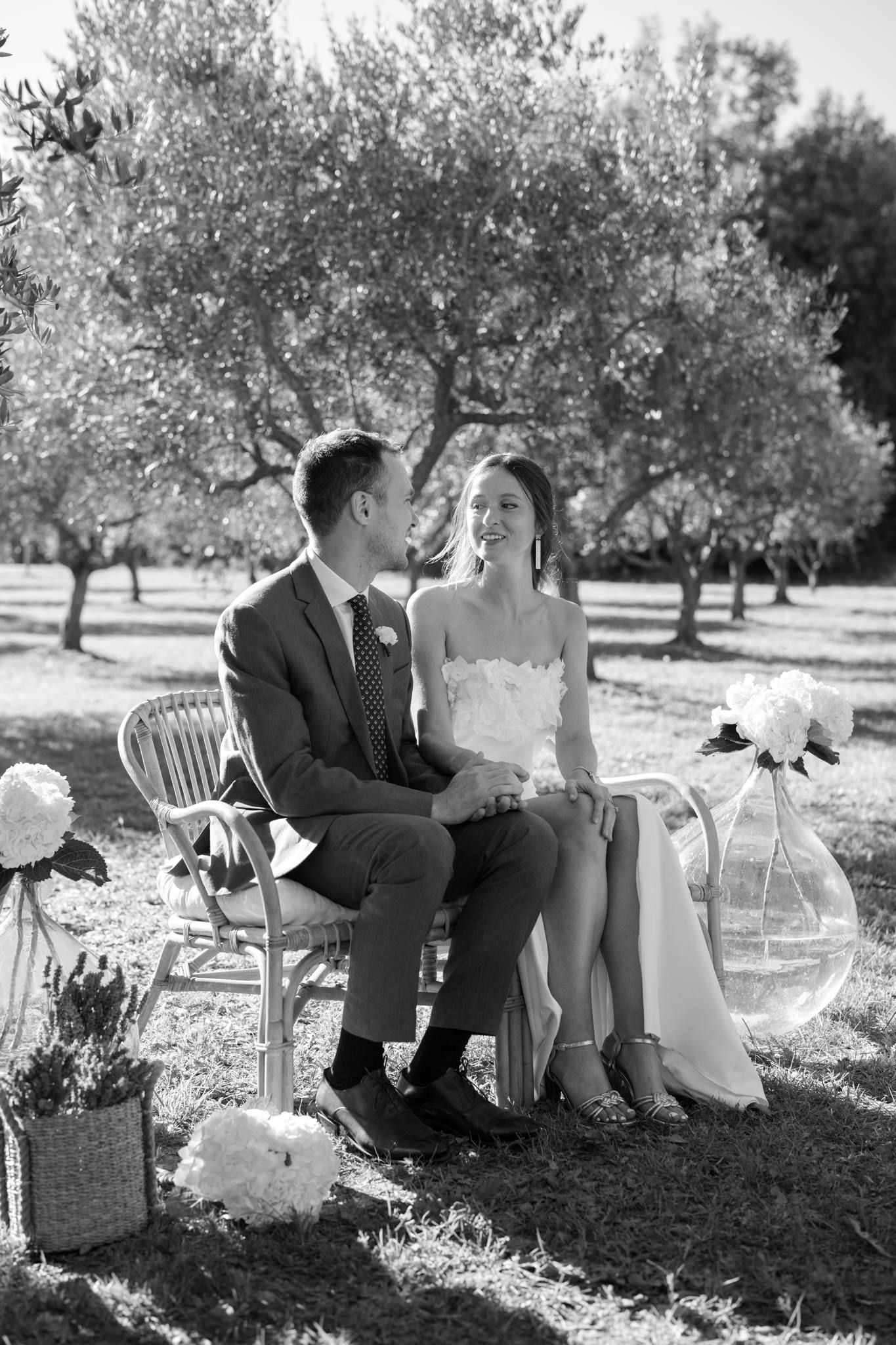 A romantic couple sits on white chairs in an olive grove during an intimate outdoor photo session.