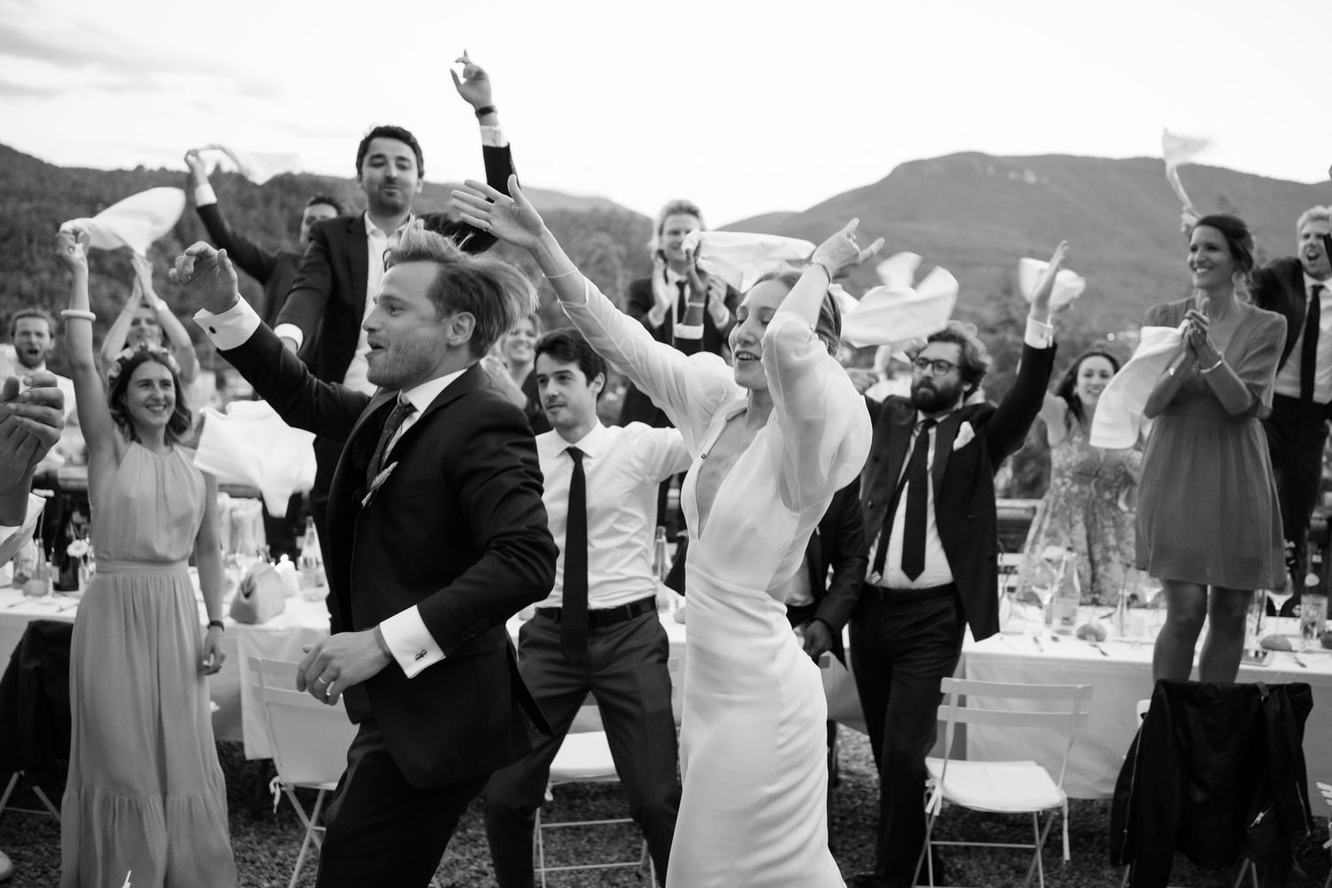 Wedding party celebrates with raised hands against mountain landscape backdrop.