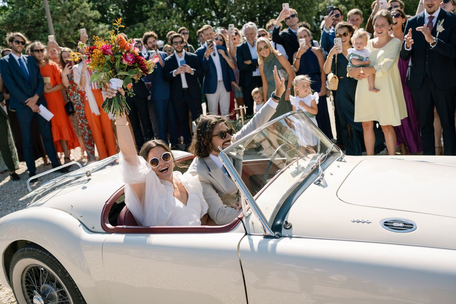 Happy couple departs wedding in vintage white convertible car surrounded by cheering guests.