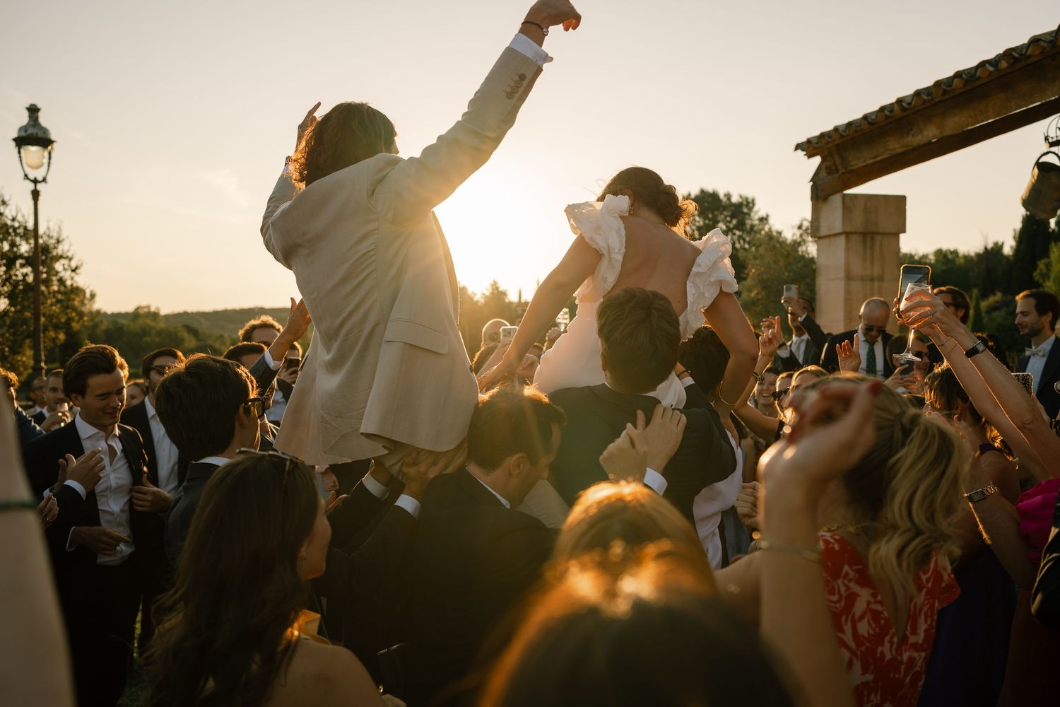 Newlyweds being lifted by crowd during sunset wedding celebration.