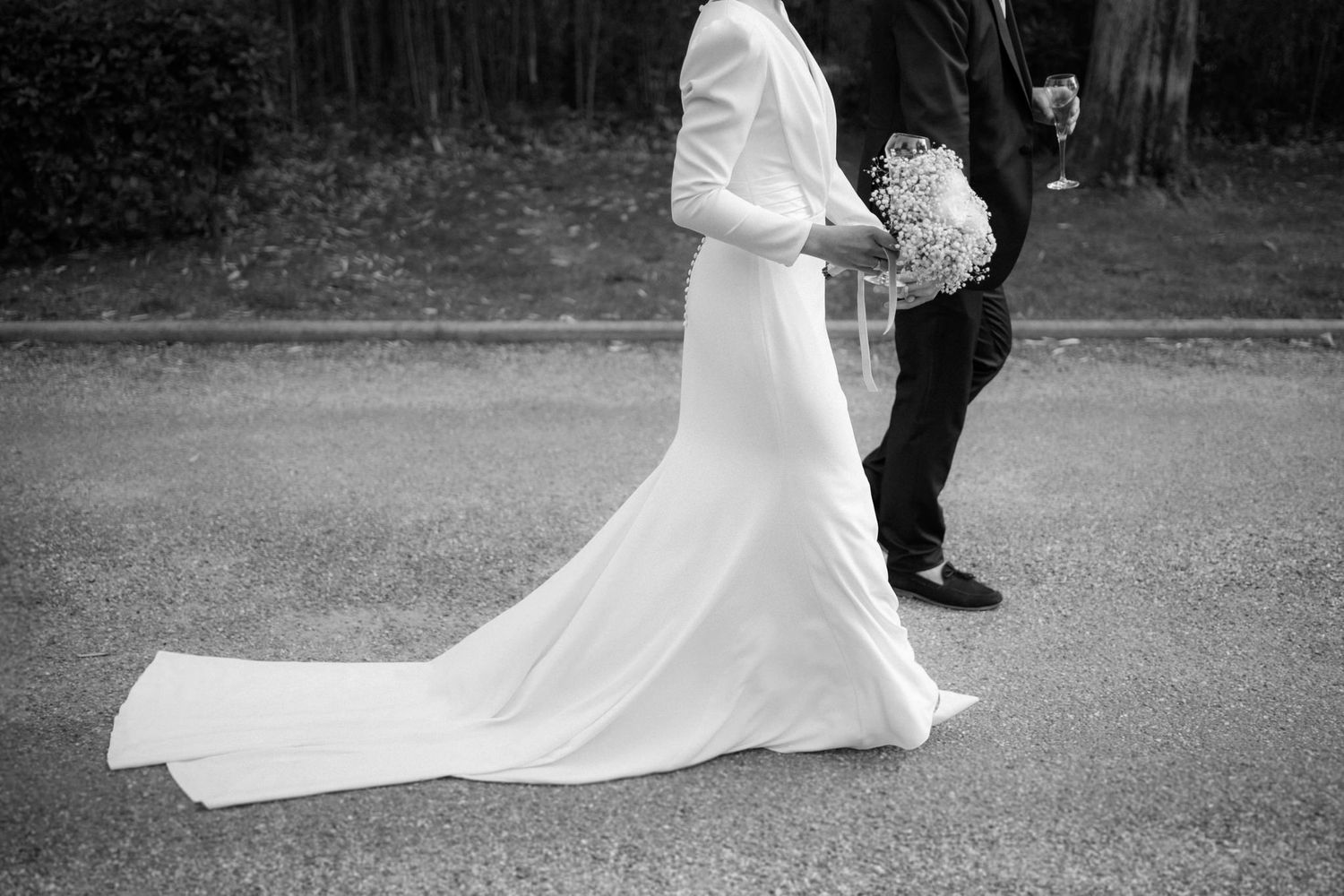 A flowing white wedding dress trails along a garden path in black and white photography.