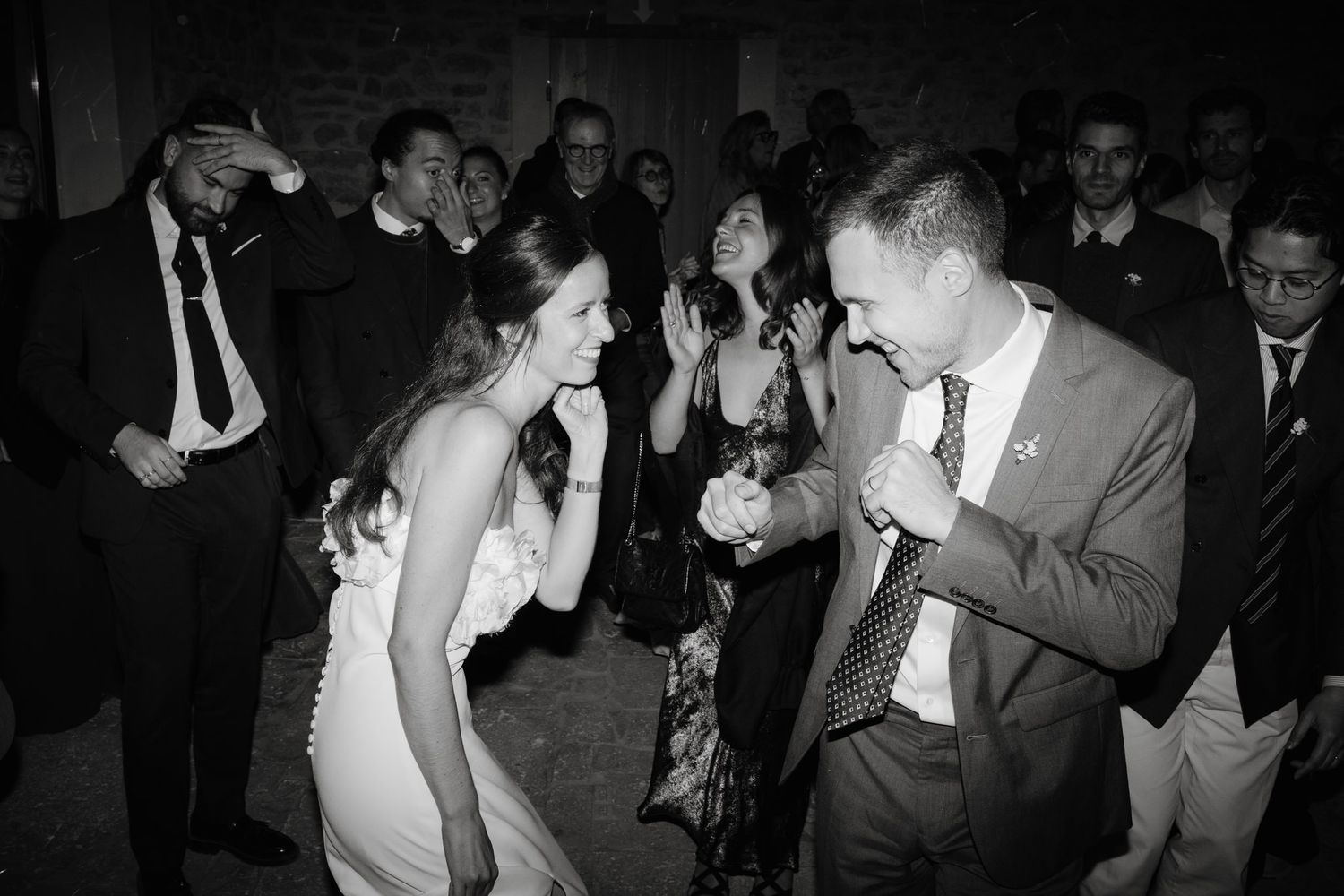 Wedding guests dance together in a dimly lit reception venue captured in black and white.