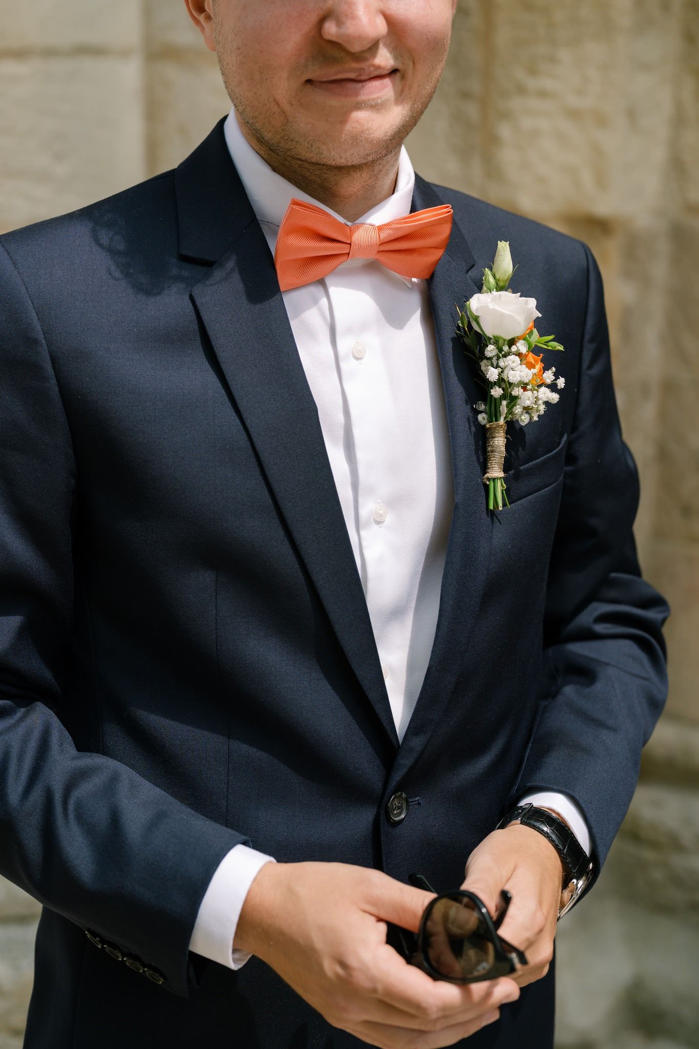 Groom in navy suit with orange bow tie and white boutonniere smiles during wedding preparations.