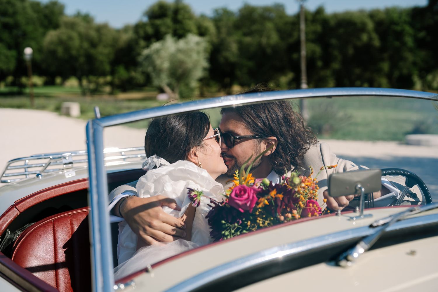 Classic convertible car parked in a sunny outdoor setting with trees and flowers in the background.