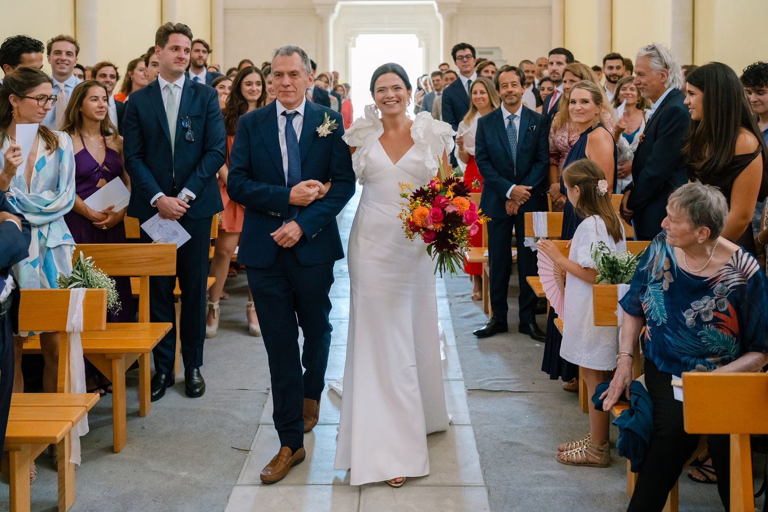 A wedding processional in a church with guests seated in wooden pews along the aisle.