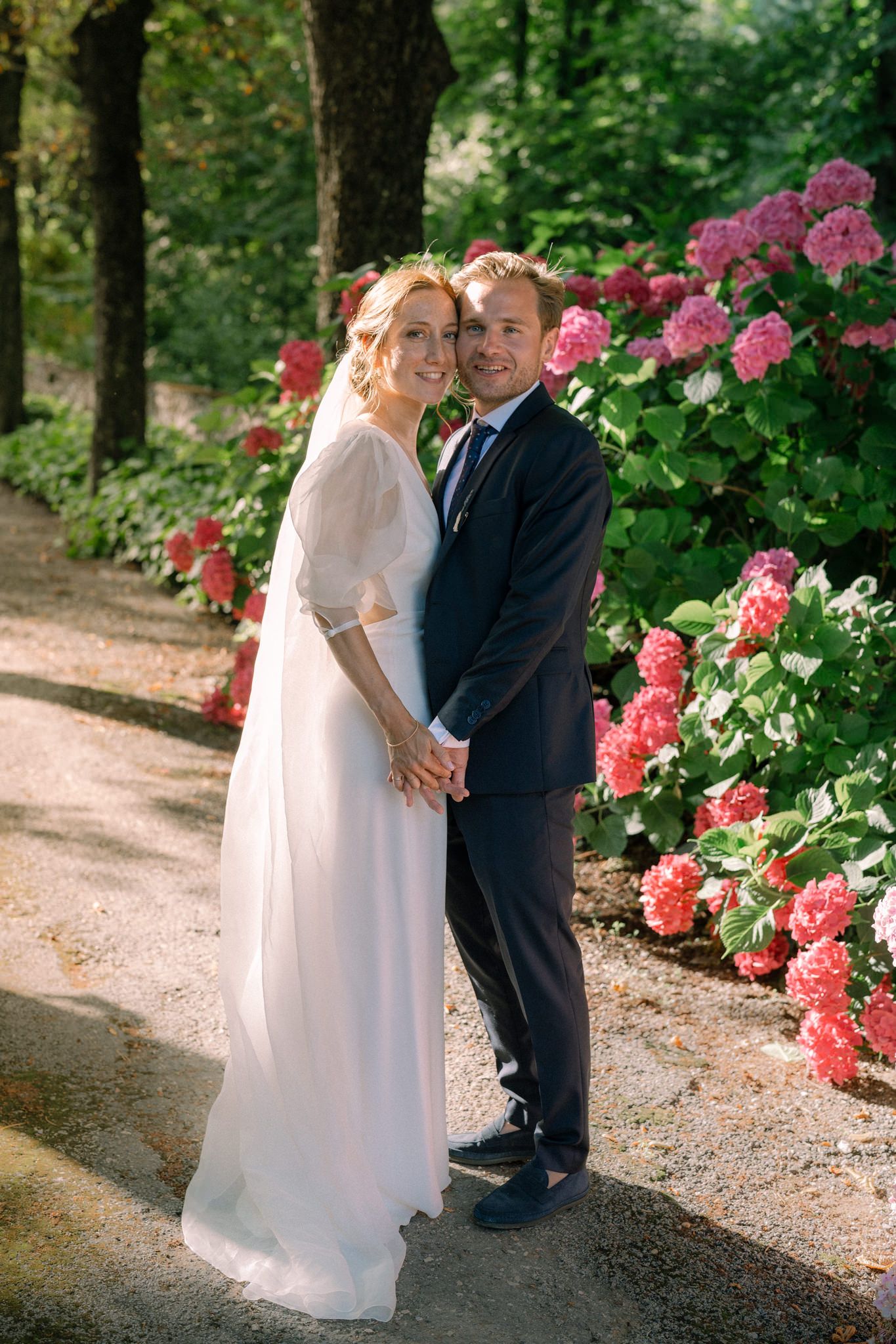 Newlyweds sharing intimate moments along a garden path with pink flowers.