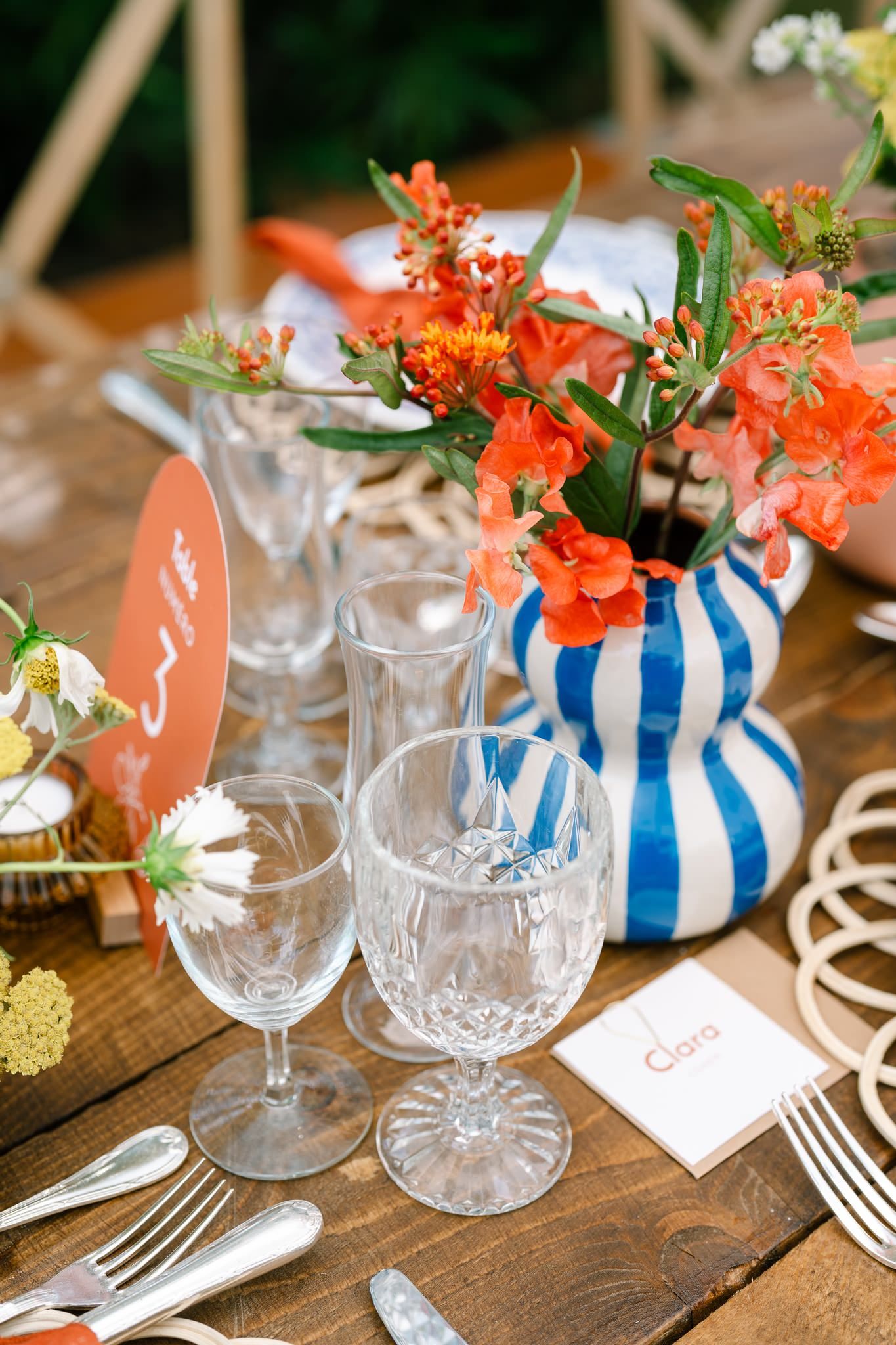 Table setting with blue striped vases, orange flowers, and crystal glassware on wooden surface.