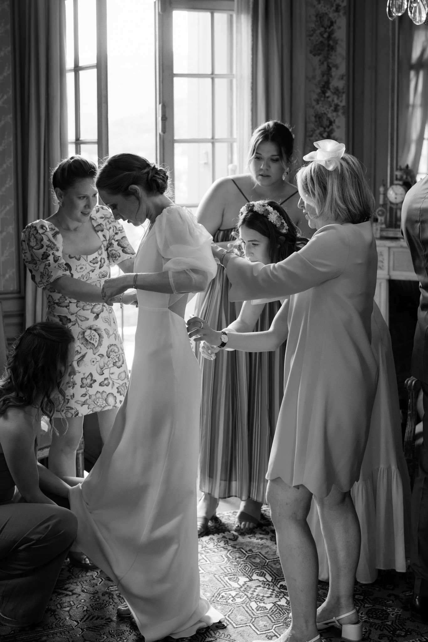 Wedding party members dance in a line inside an elegant room with large windows.