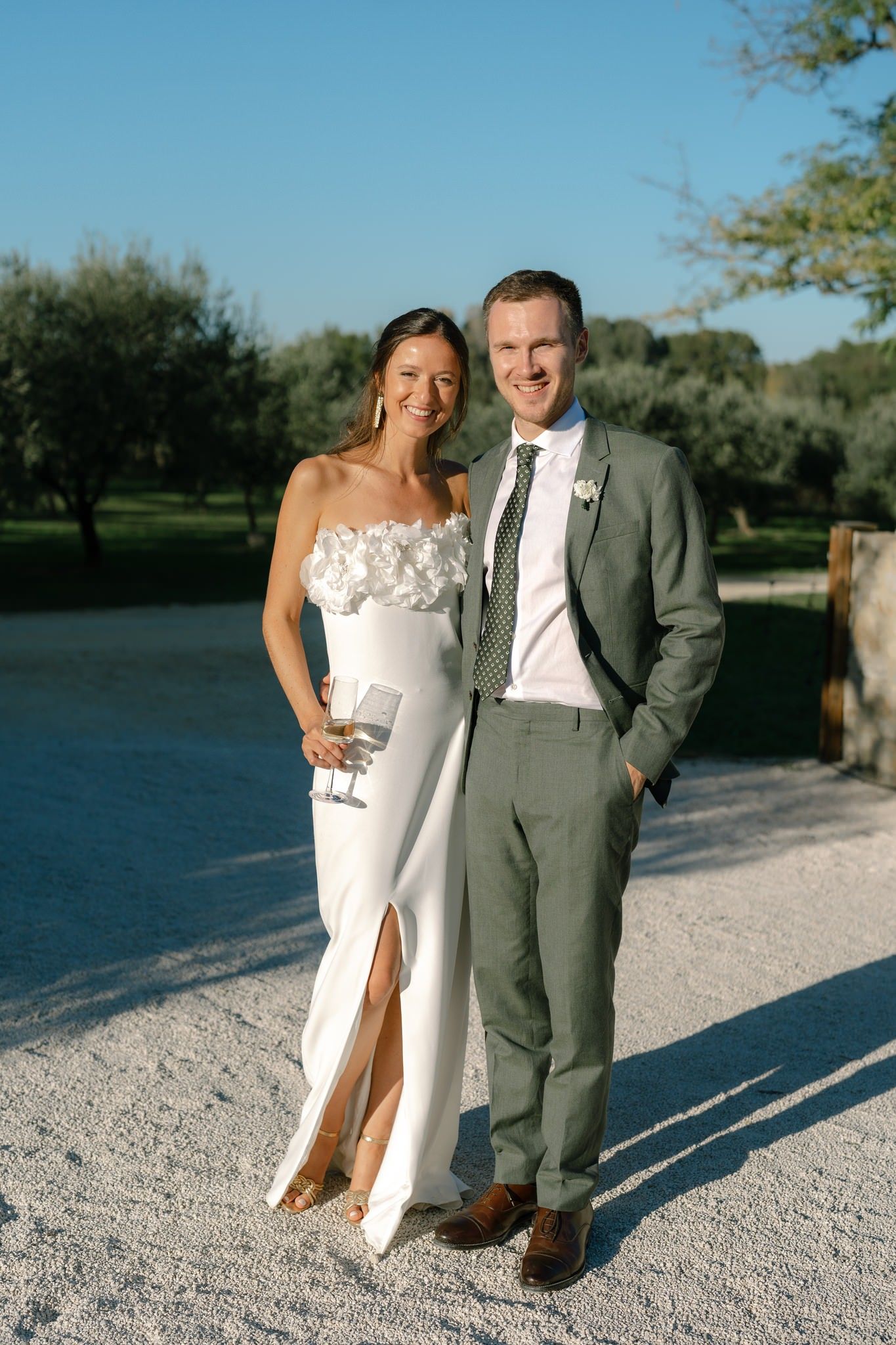 A couple poses together on a white gravel path near a lake in wedding attire.