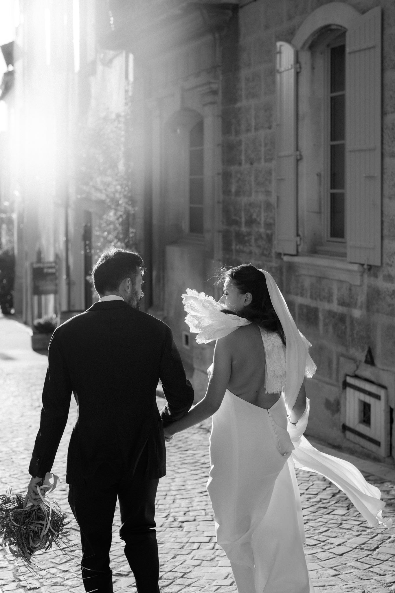 A couple walks hand in hand down a sunlit cobblestone street.