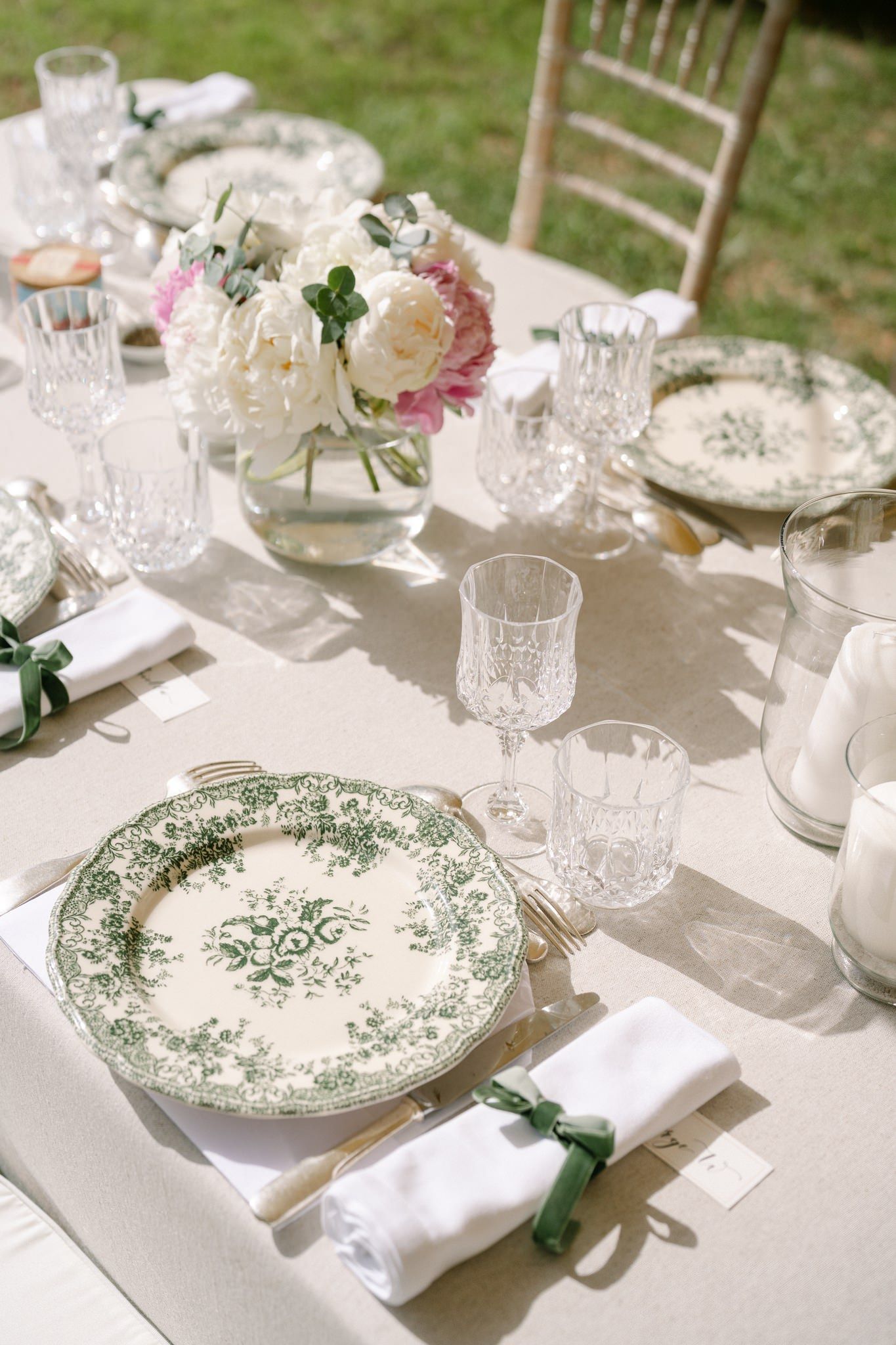 Elegant table settings with vintage patterned plates, crystal glasses and white linens at an outdoor reception.