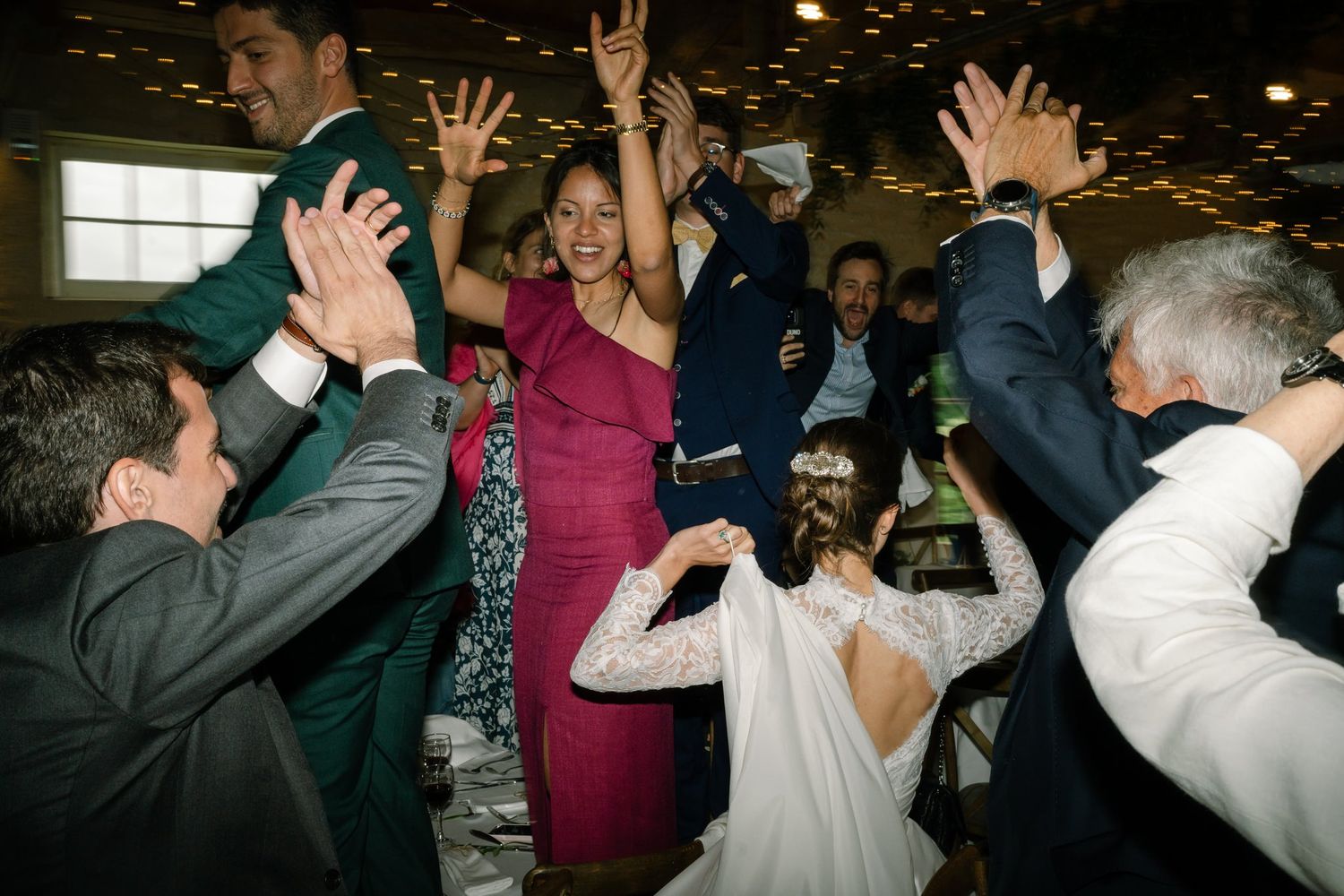 Wedding reception dance party with hands raised in celebration under dim lighting.