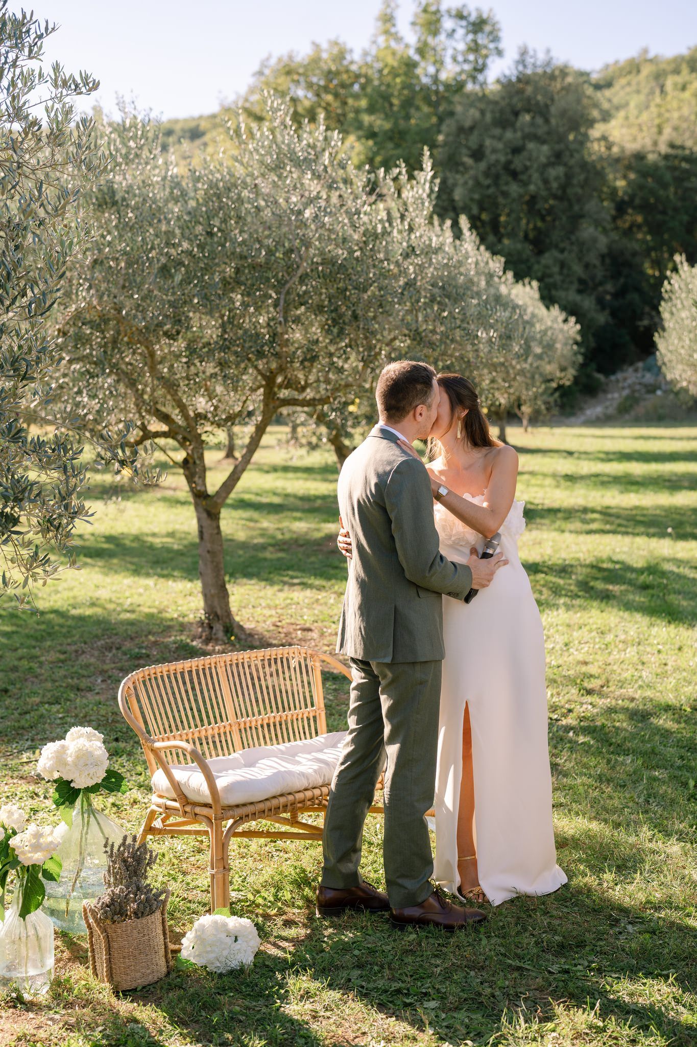 A couple shares an intimate moment by a wooden bench in an olive grove.