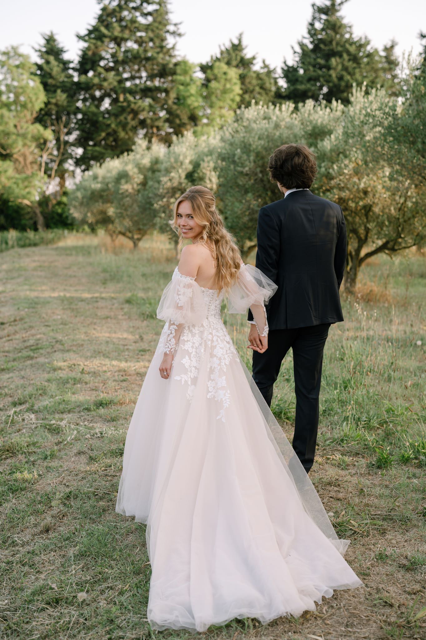 Bride in flowing lace gown walking through an olive grove with groom.