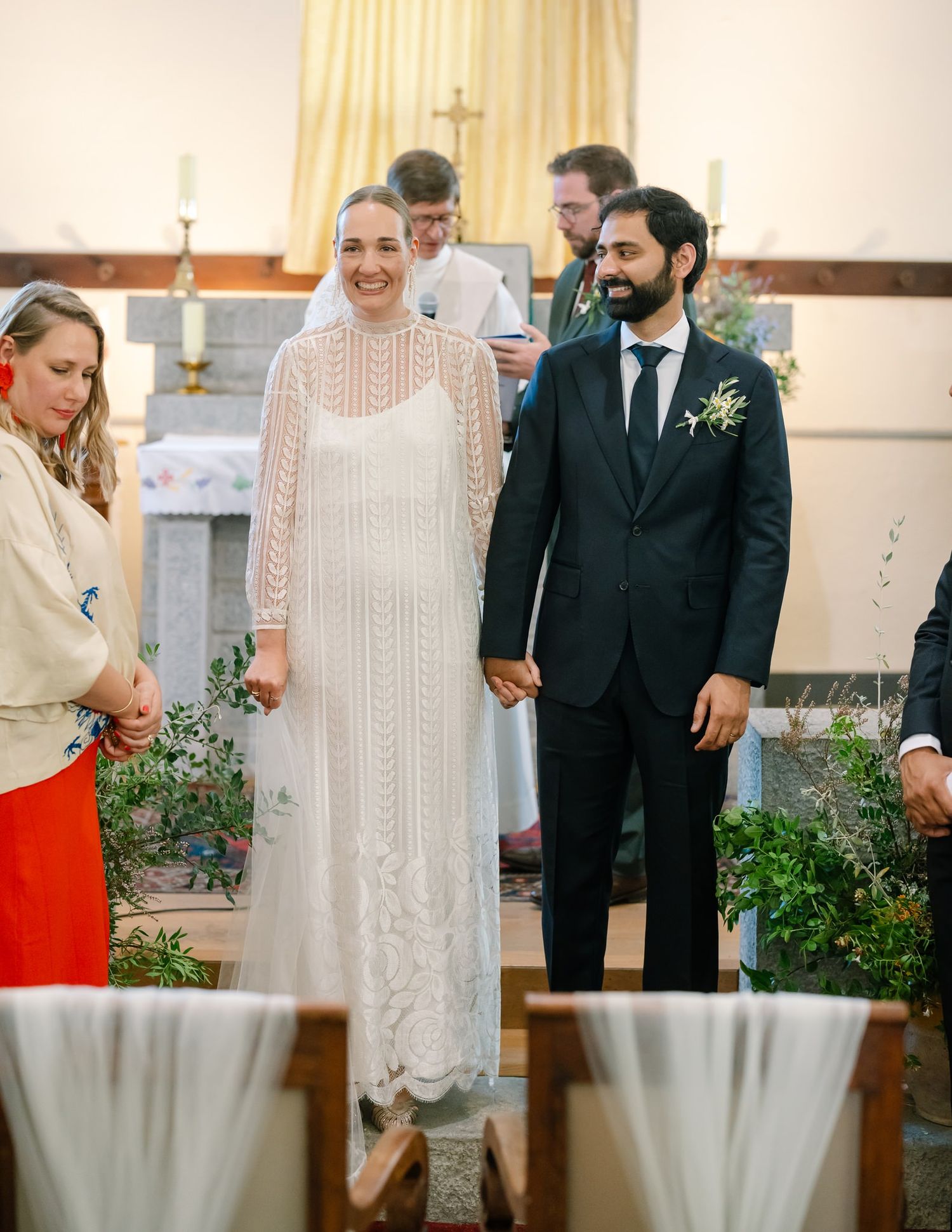 Wedding ceremony participants stand in church with decorative plants and white decor.