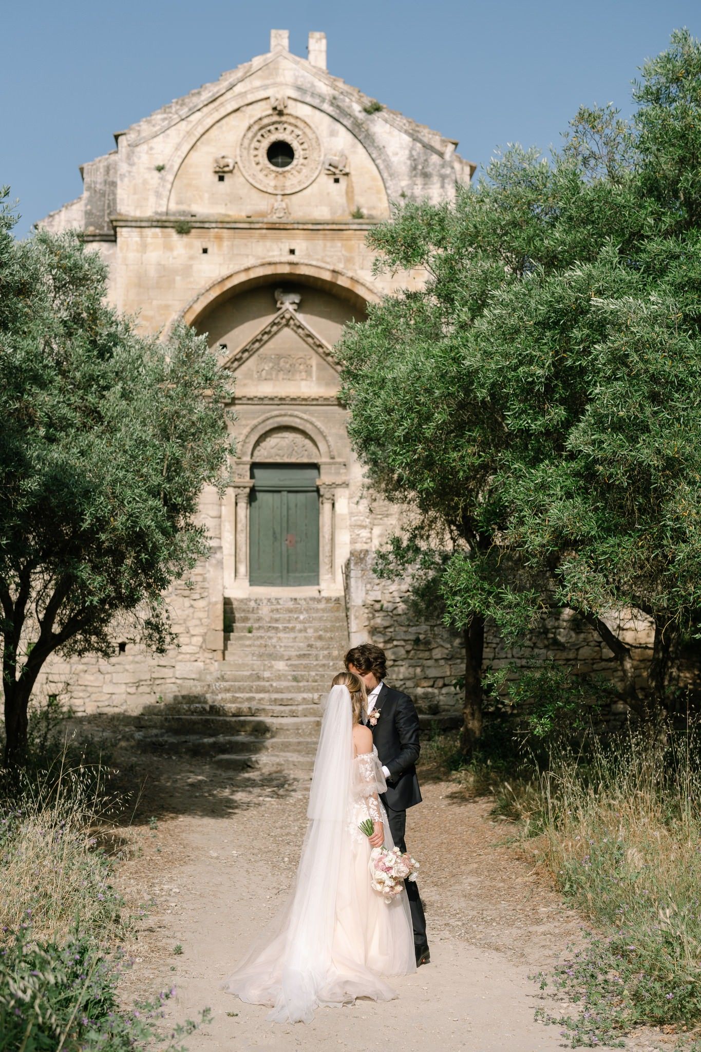 Couple taking wedding photos in front of a rustic stone chapel with olive trees.