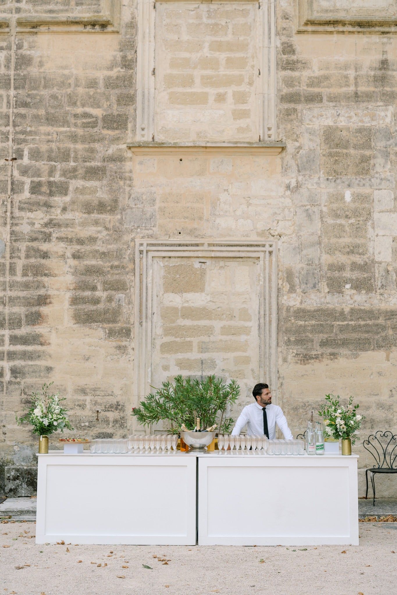 White modern bar setup with greenery against a stone wall at an elegant outdoor venue.