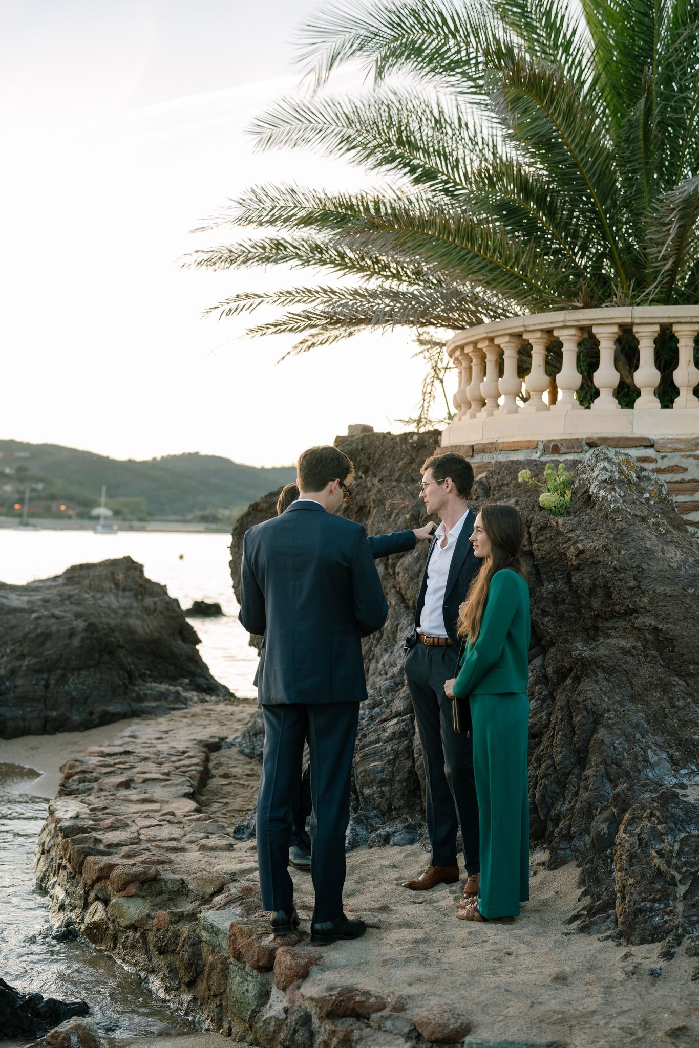 Wedding guests gather on rocky coastal overlook with palm trees and ocean view.