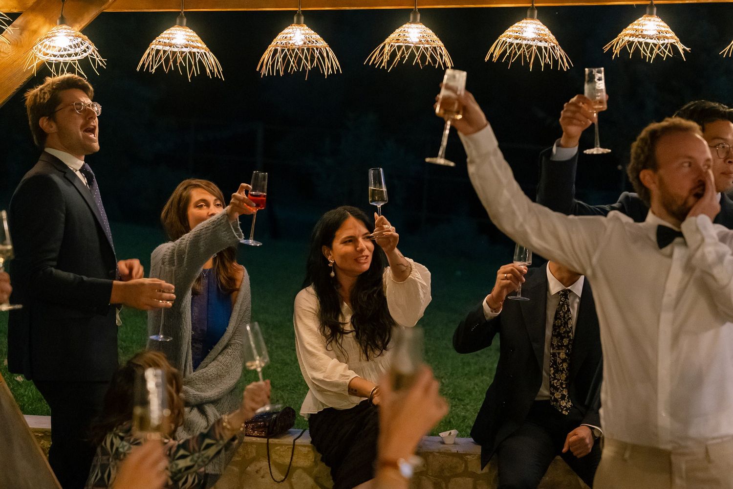 Wedding celebration at night with string lights and guests raising glasses in toast.