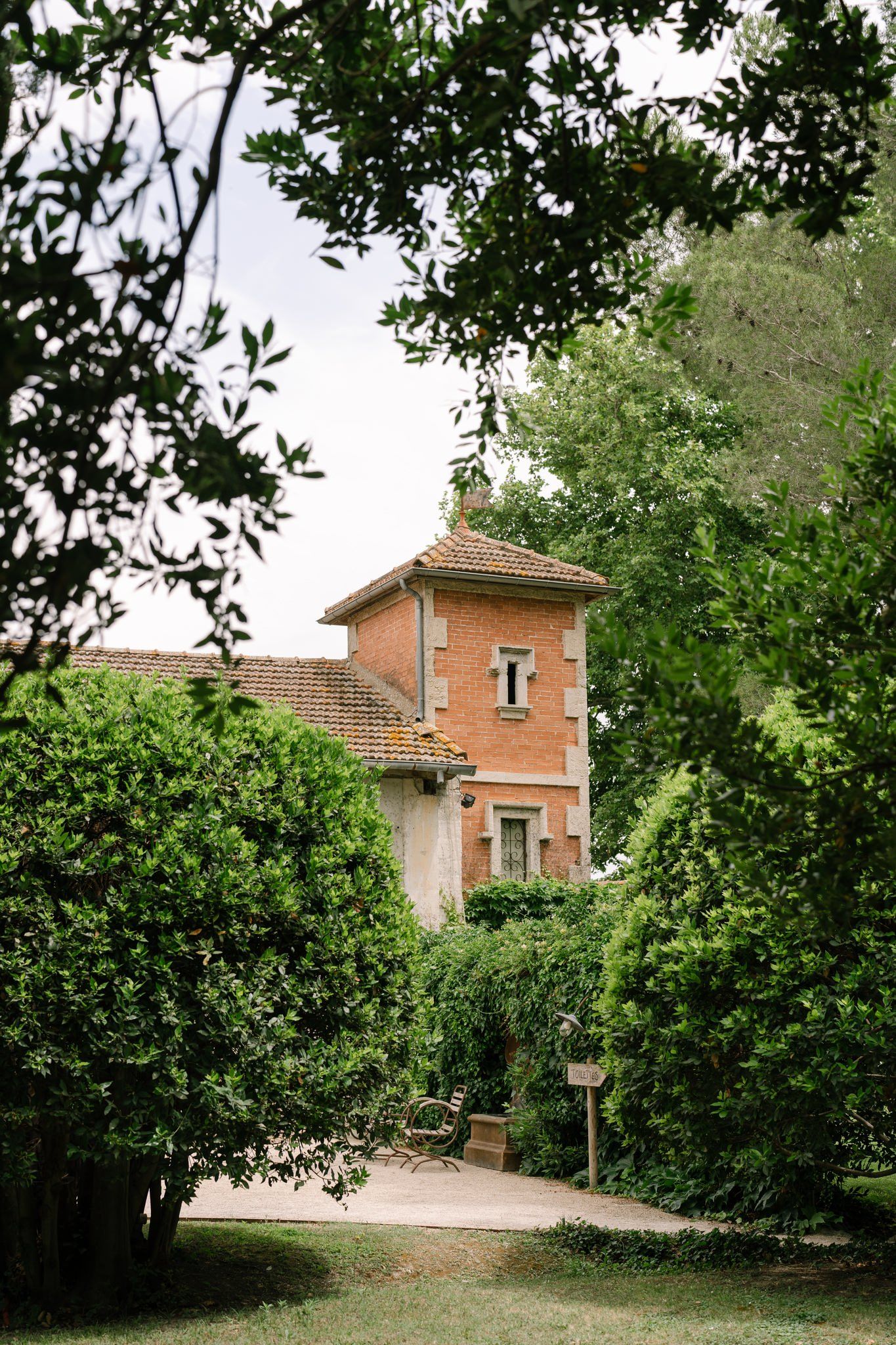 A brick villa with terracotta roof peeking through lush green trees and foliage.