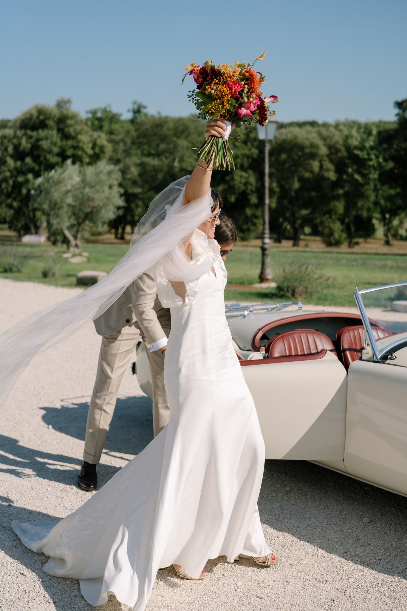 A couple poses with a vintage car on a sunny day with flowing white dress and veil.
