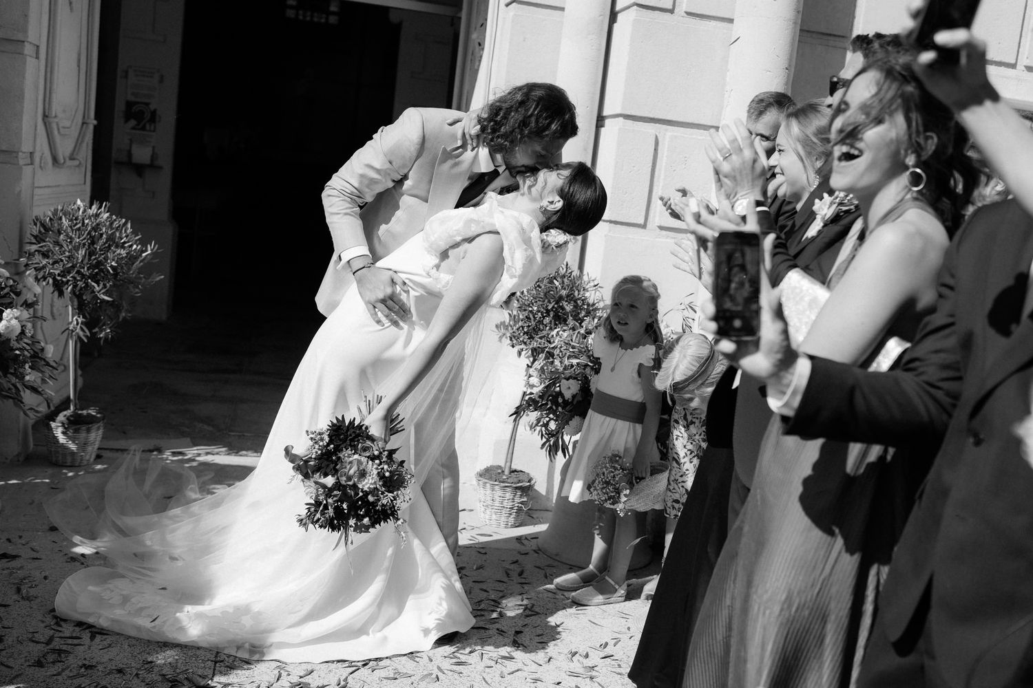 Black and white photo of a romantic wedding moment surrounded by guests outside a historic venue.