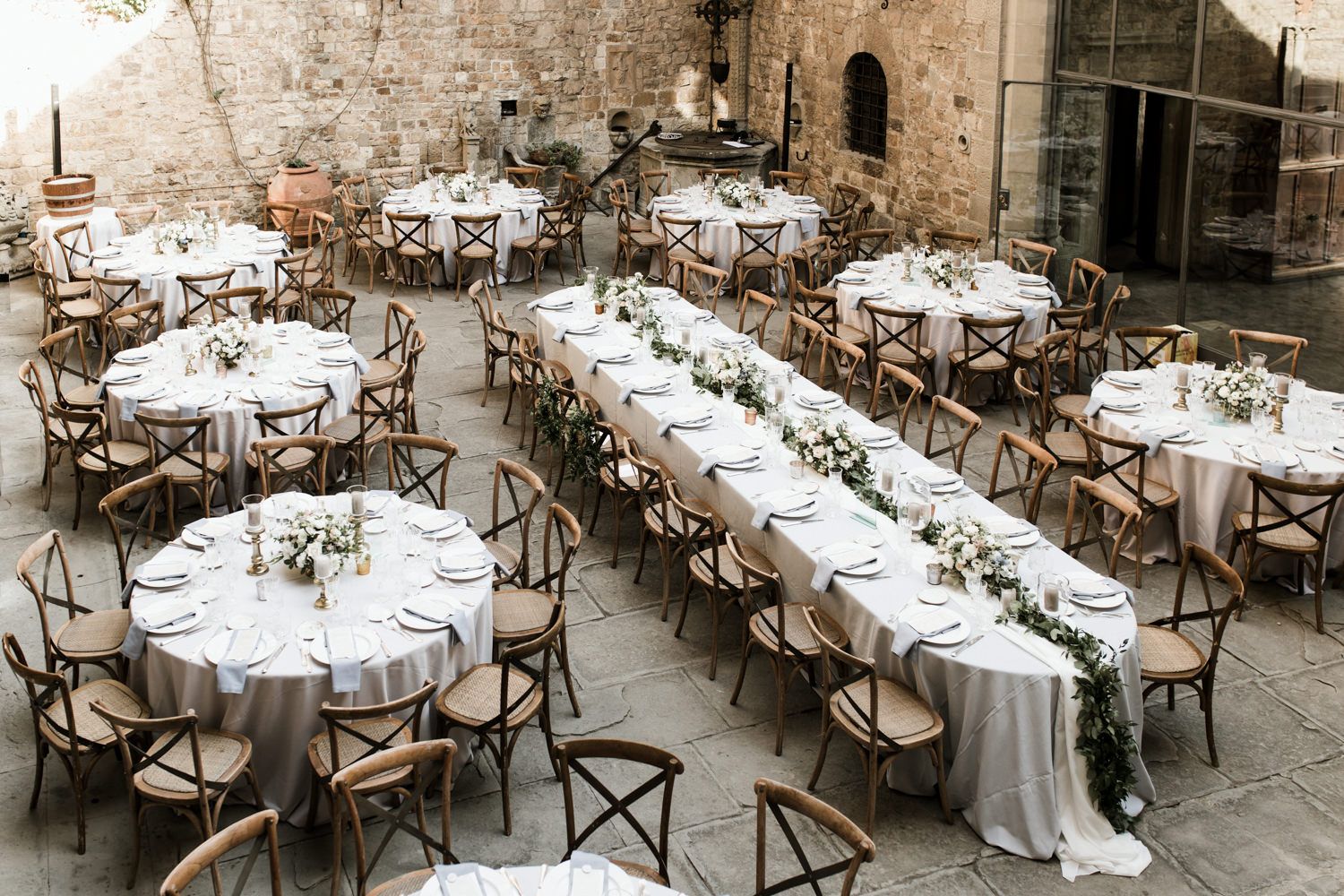 Round dining tables with white linens and cross-back chairs set up in a historic courtyard.