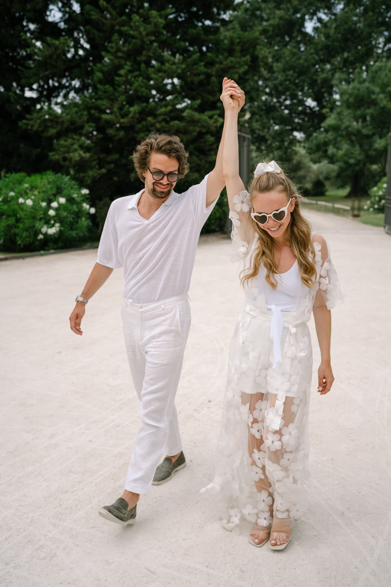 A couple in all white outfits walking together on a path surrounded by greenery.