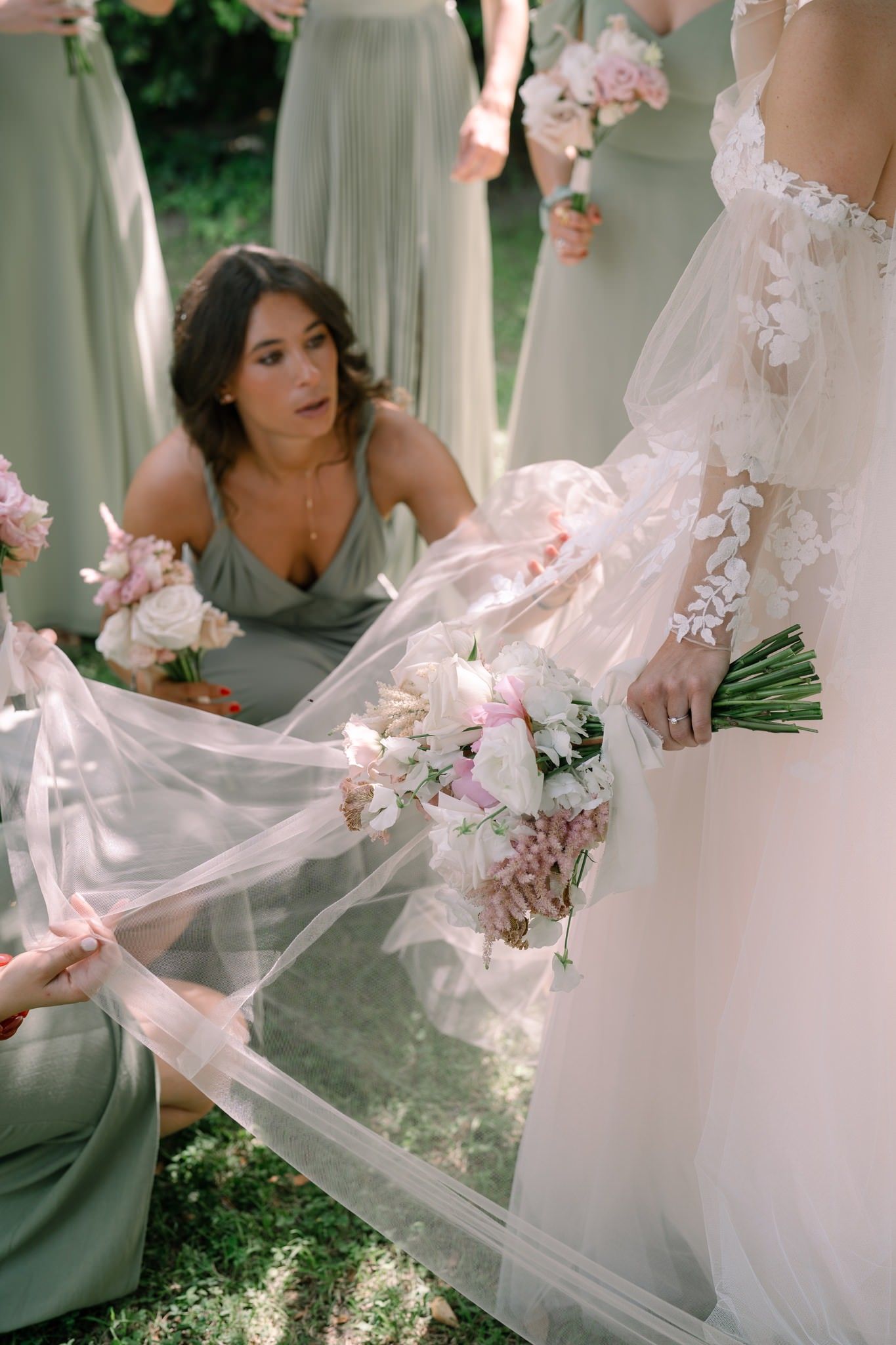 Wedding party arranging the bride's flowing white dress train and veil outdoors.
