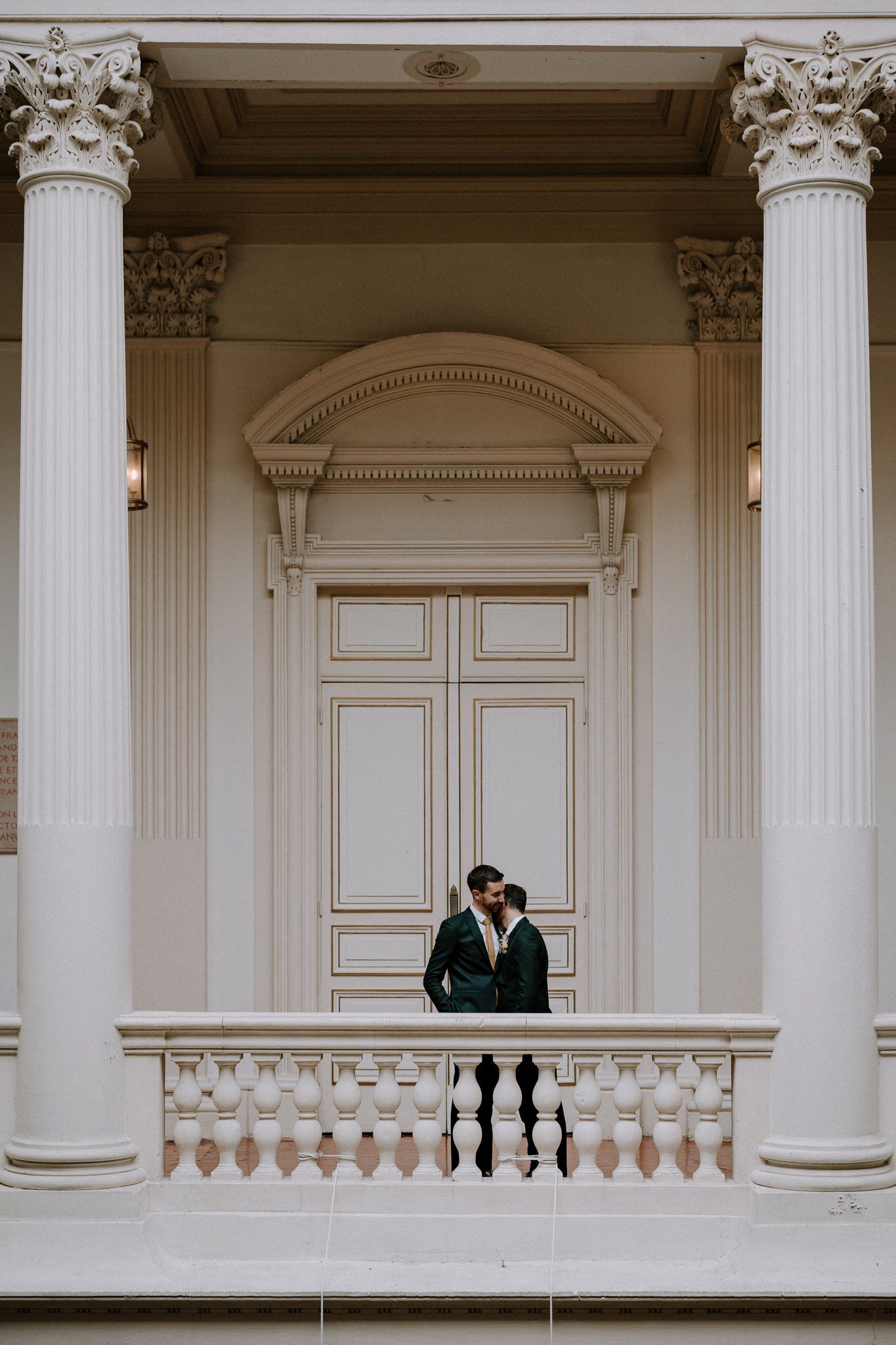 Two figures stand on an elegant white columned balcony in formal attire.