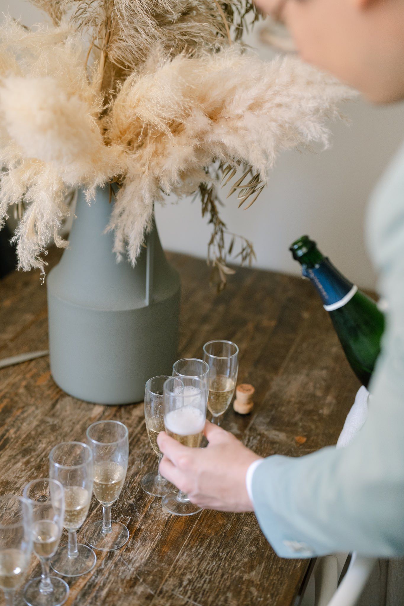 Champagne being poured into glasses with pampas grass decoration on wooden surface.