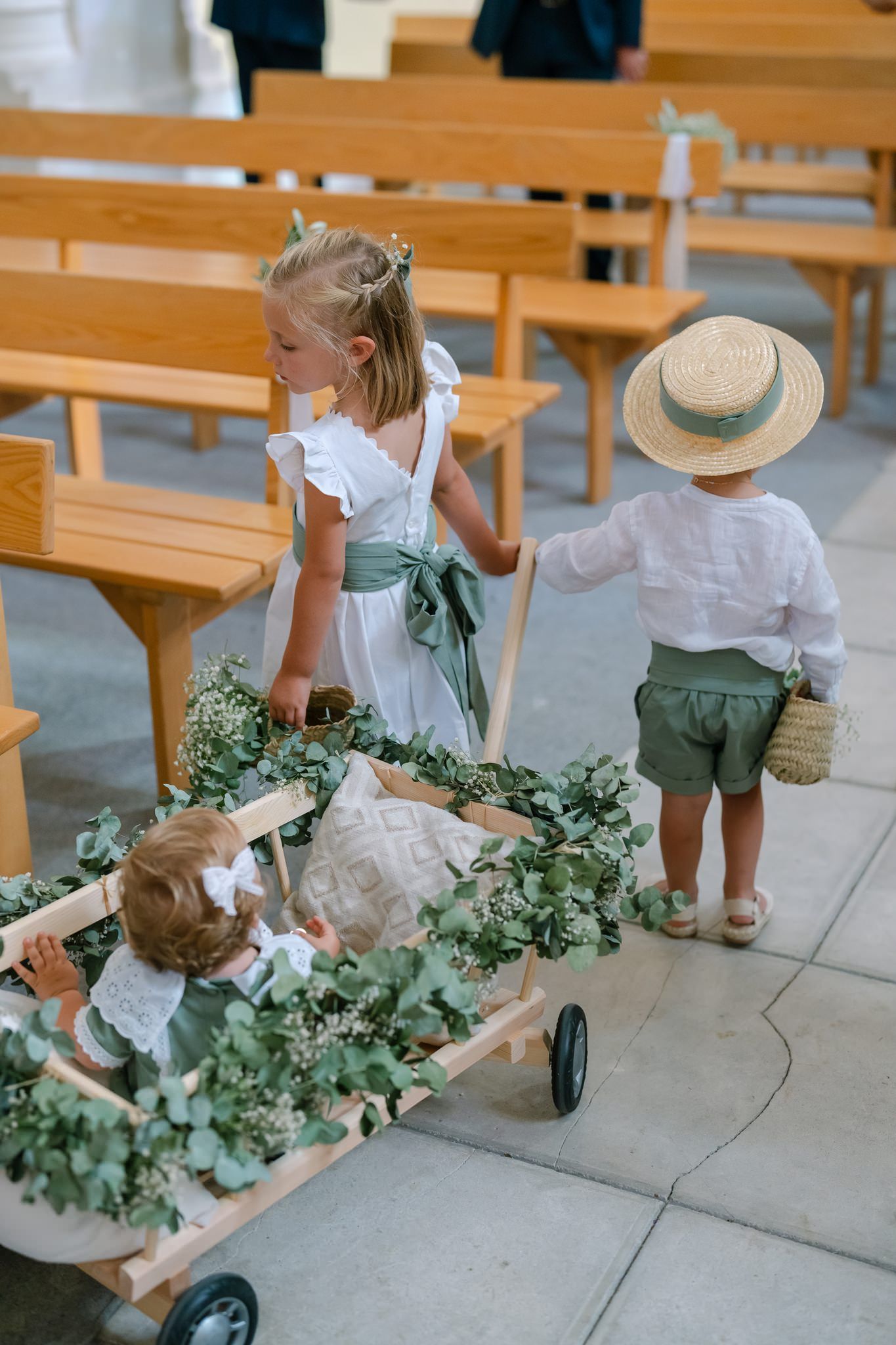 Children in summer attire pulling decorated wagons down a church aisle.