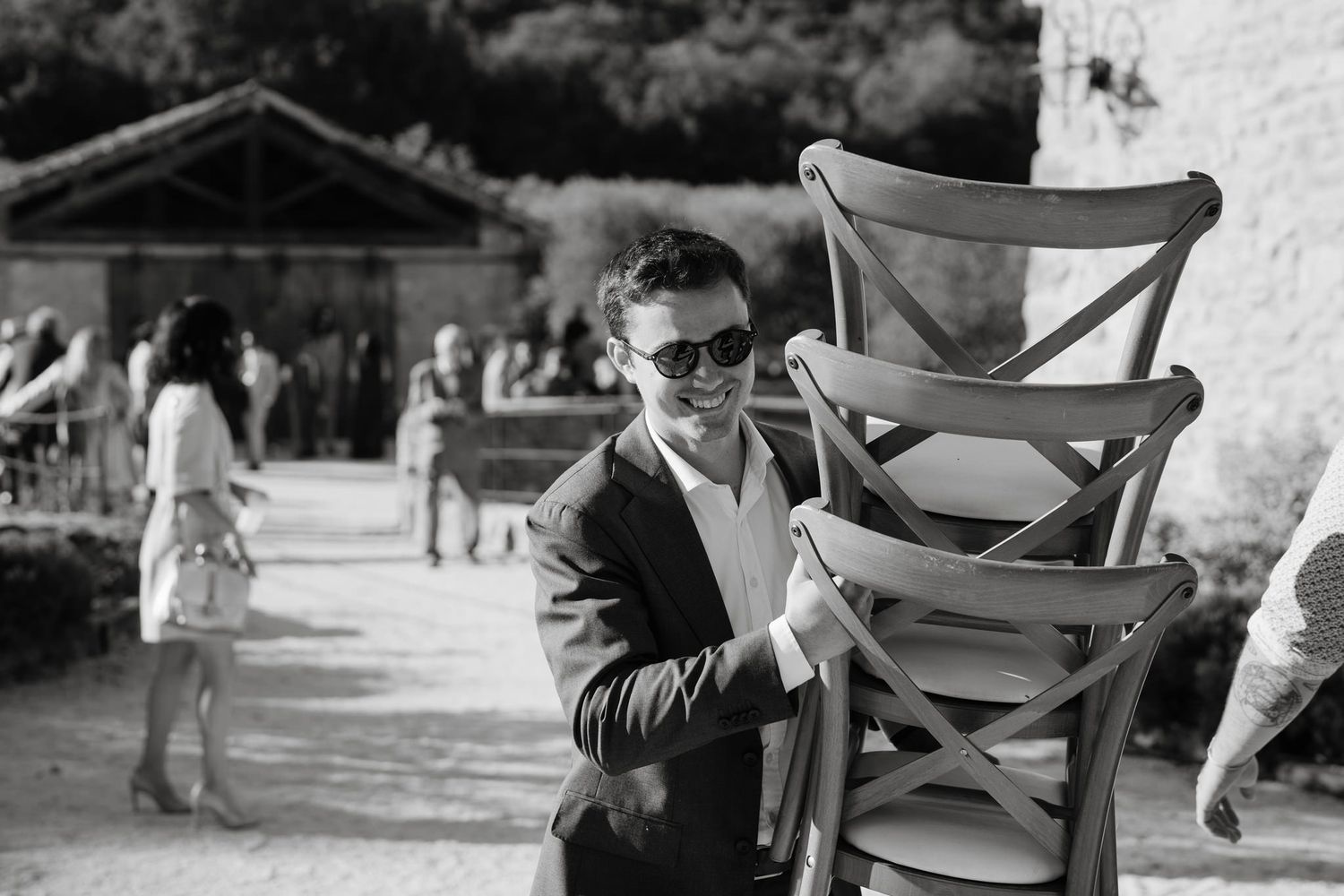 Person carrying stacked wooden chairs at an outdoor venue.