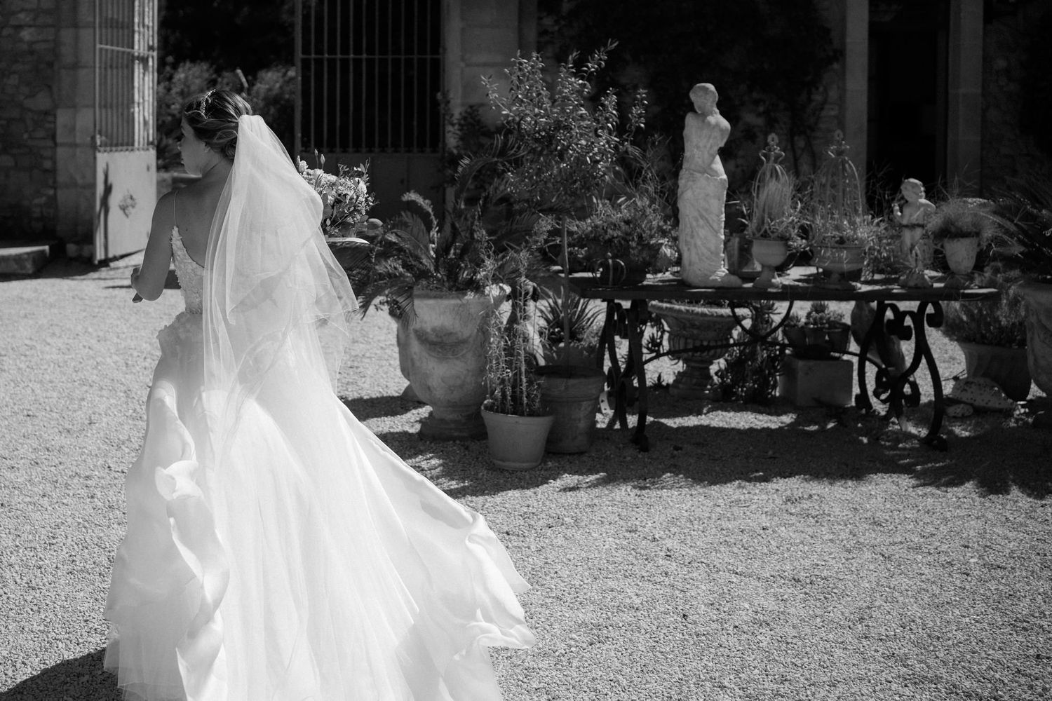 Elegant bride in flowing white dress walking through a stone courtyard on her wedding day.