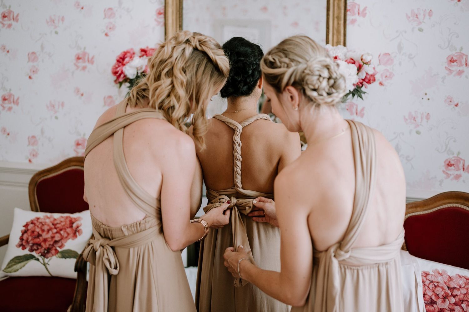 Elegant wedding preparation scene with bridesmaids in neutral dresses against floral wallpaper.