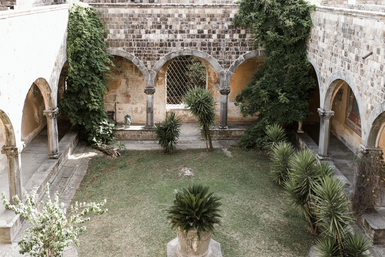 Stone archways and columns line a rustic courtyard garden with lush greenery and palm trees.