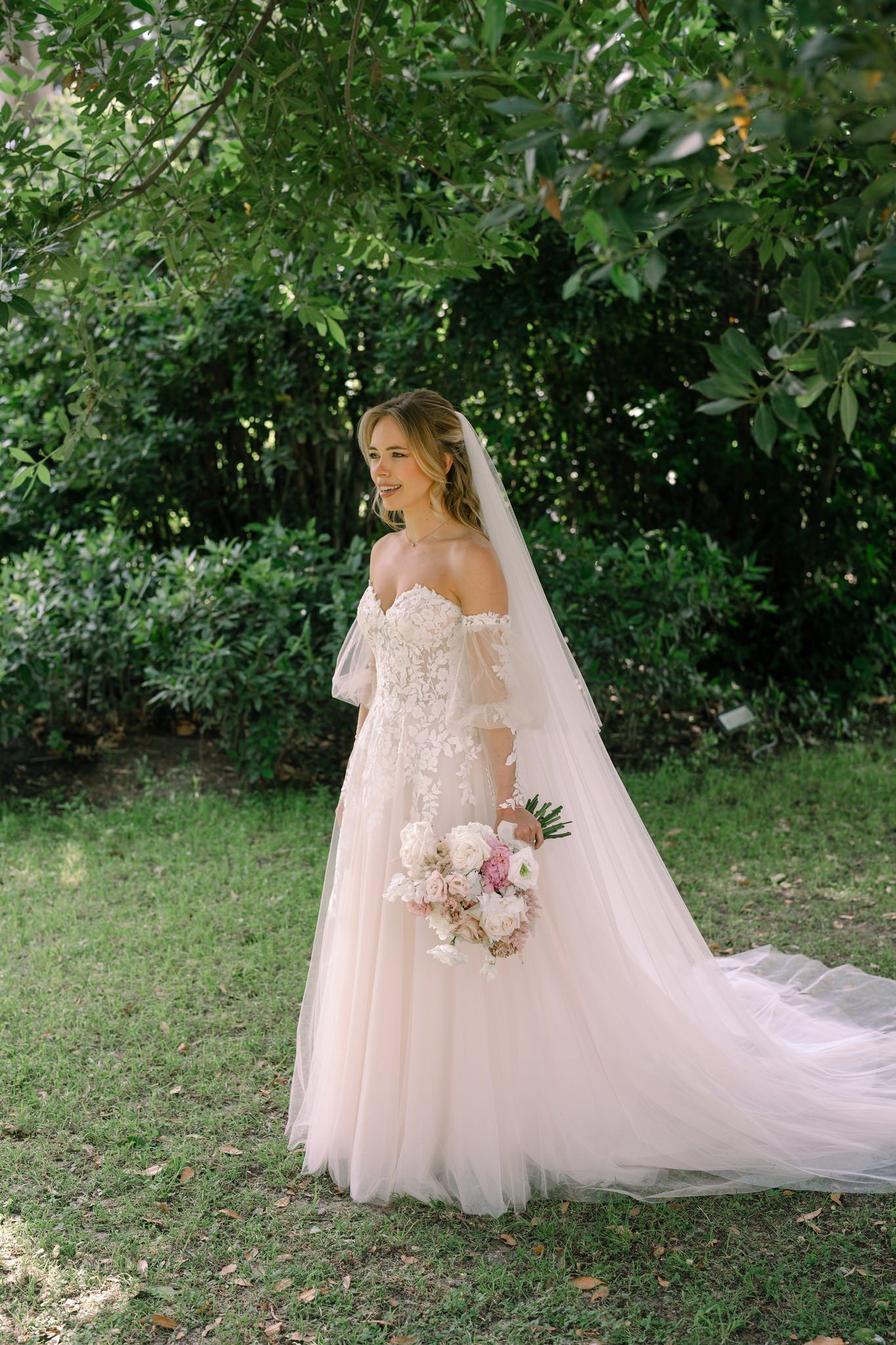 Wedding dress with off-shoulder design photographed in a garden setting with a flowing veil.