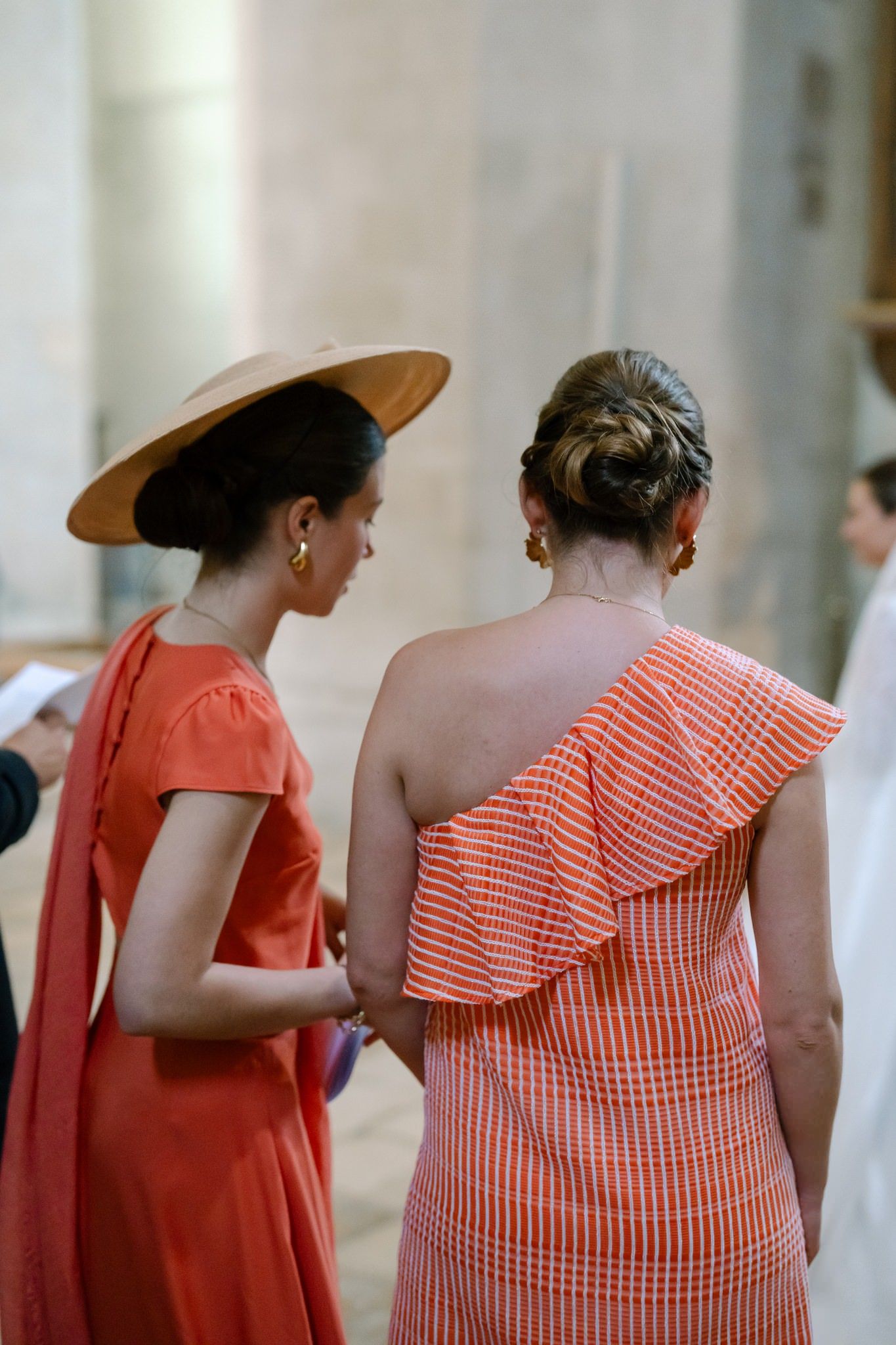 Two wedding guests in coral dresses chat during reception.