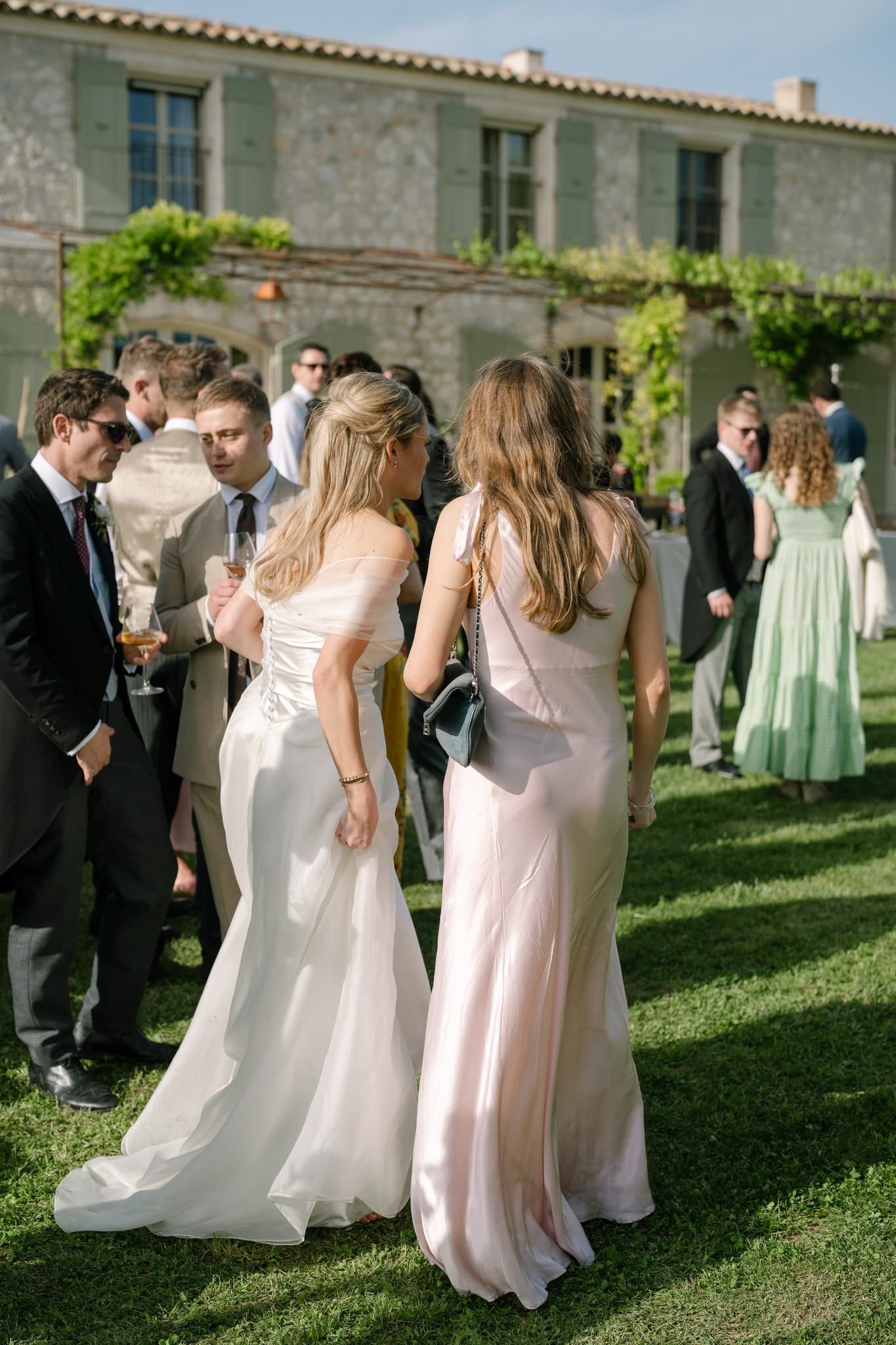 Wedding guests mingle on lawn in formal attire outside stone villa.