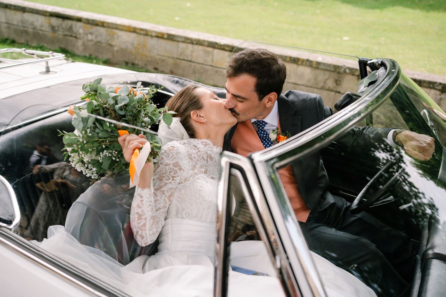 Newlyweds riding in a classic white convertible car through scenic countryside.