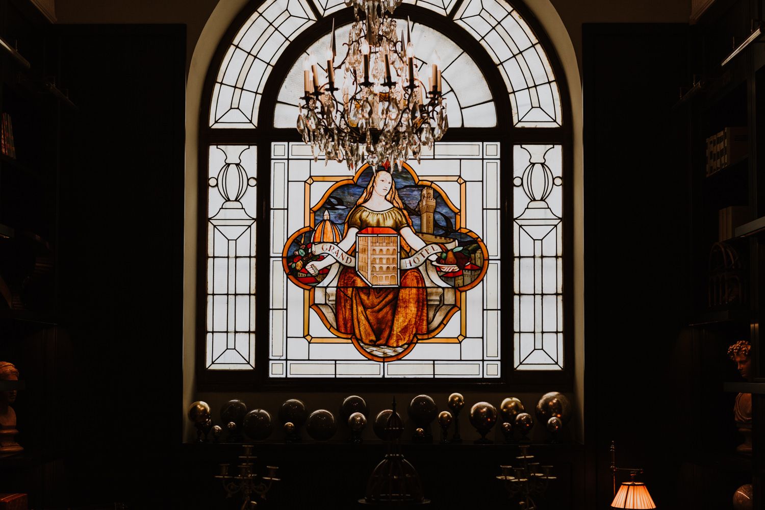 Ornate stained glass window with religious artwork illuminated by a grand crystal chandelier in a church setting.