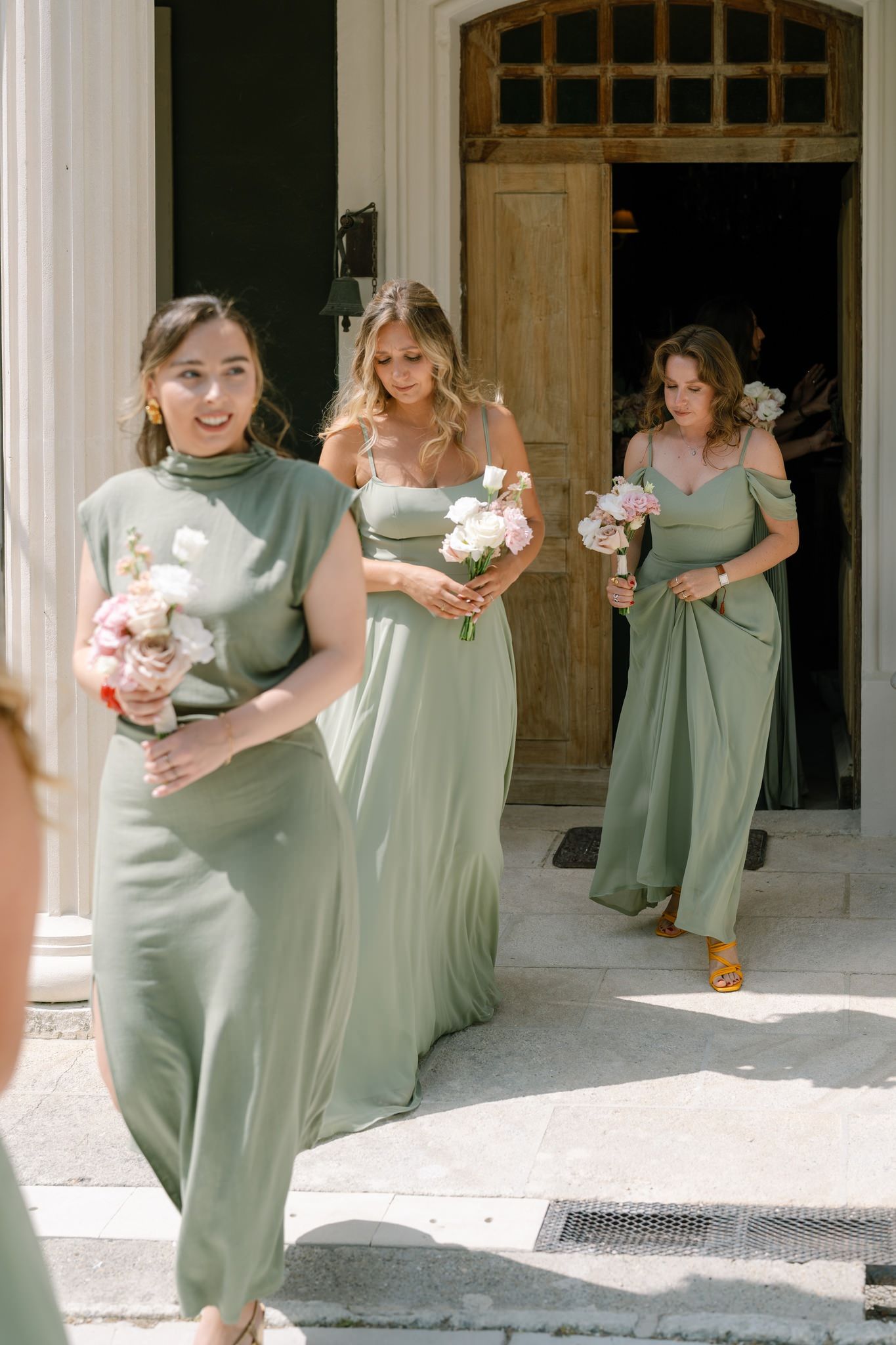 Bridesmaids in sage green dresses walking with bouquets outside a historic building.