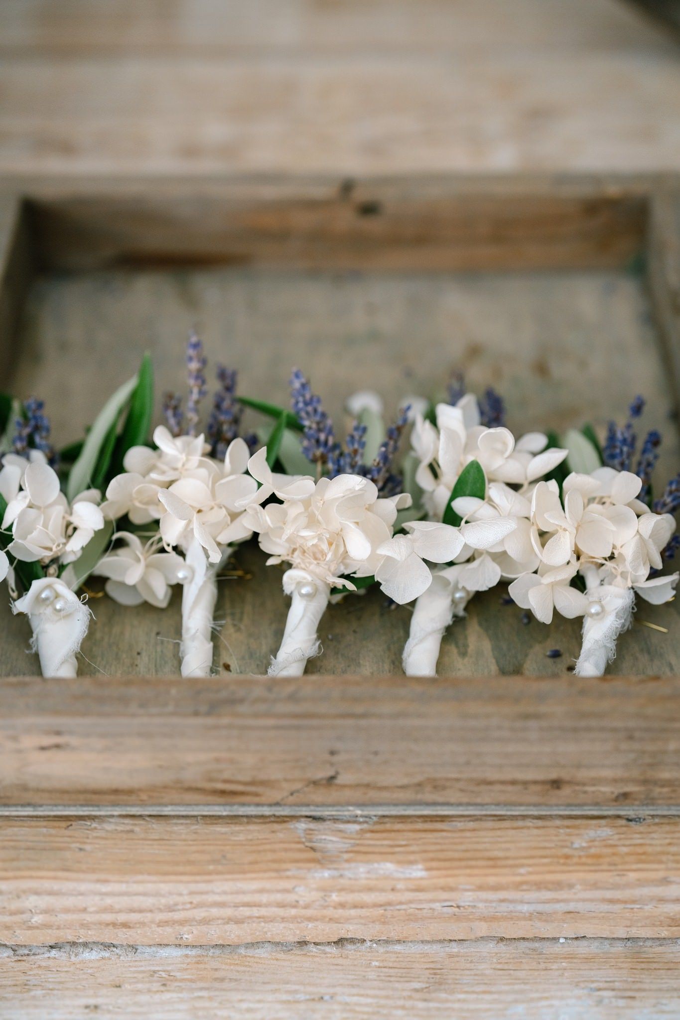 Row of delicate white floral boutonnieres with purple accents arranged on rustic wooden surface.